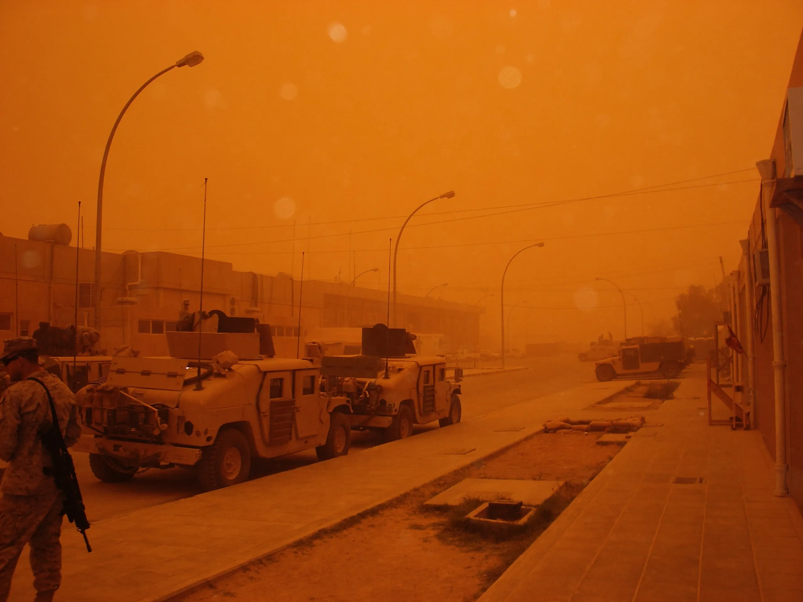 Military vehicles line a street under orange dust-filled skies, with a person partly visible at the left edge.