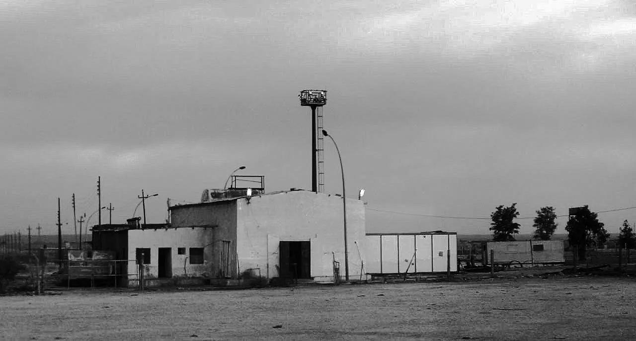 Black-and-white photograph of a small utilitarian building with a tall light tower and utility poles.