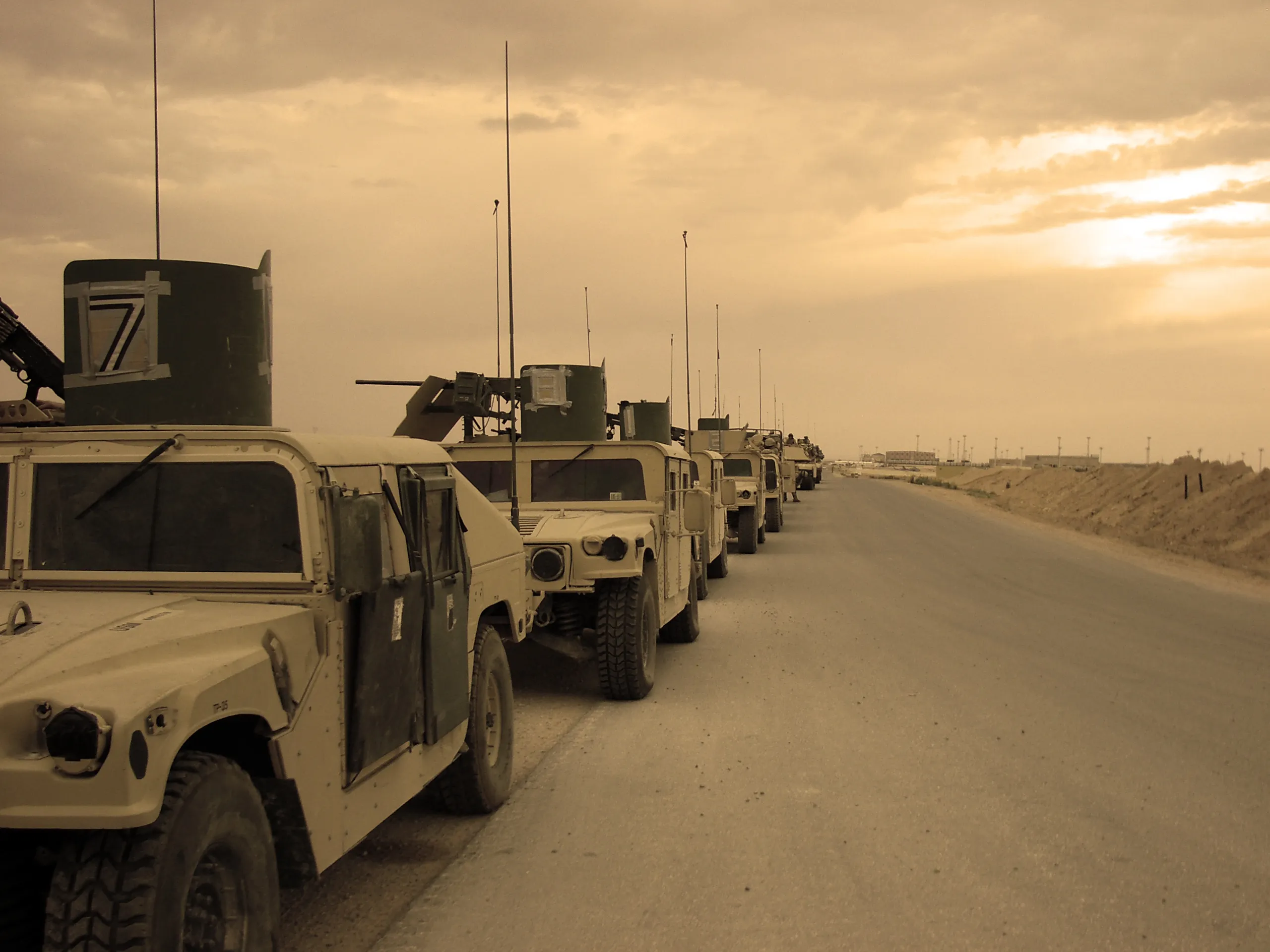 Several military vehicles parked in a line on the shoulder of a long road under a yellow-gray sky.