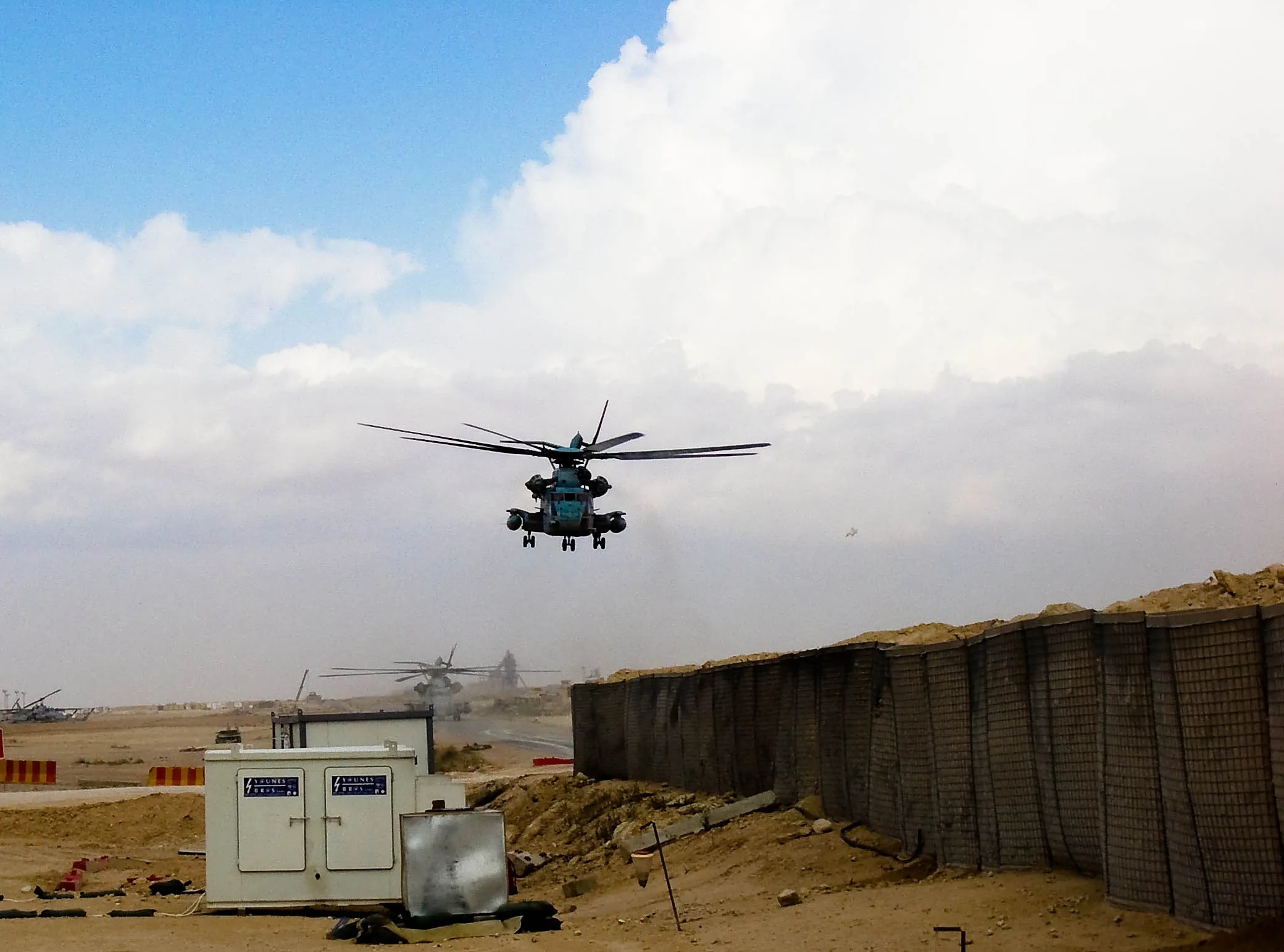 A military helicopter hovers above a dusty landscape beside tall protective barriers, with another helicopter in the distance.