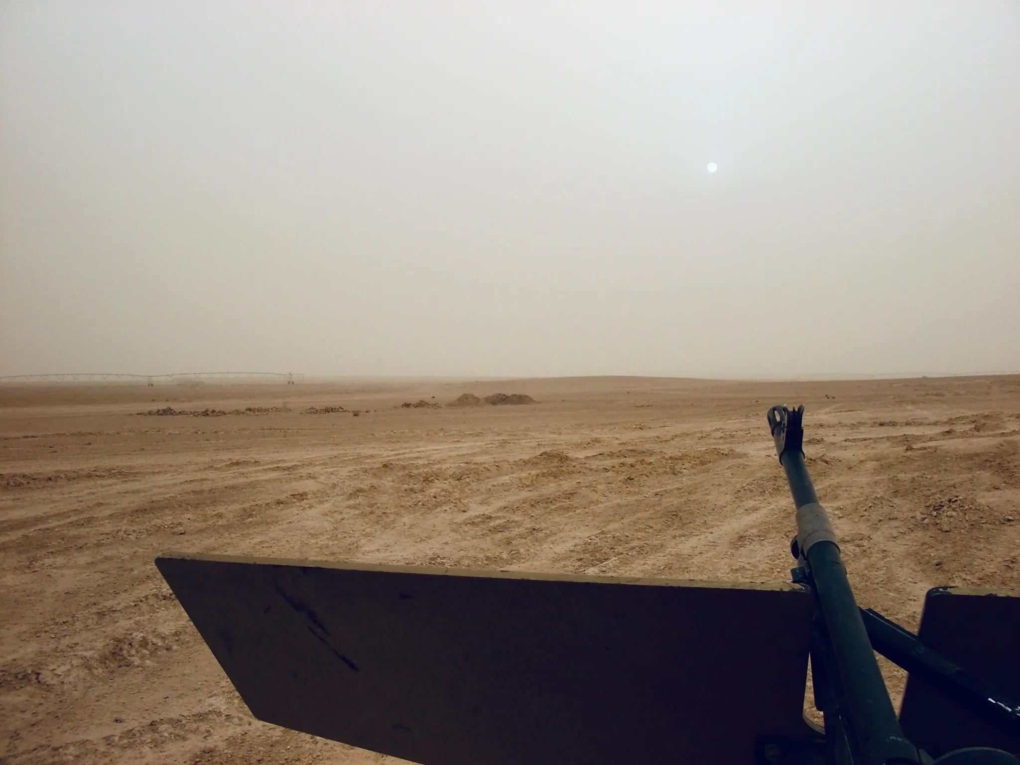 View from a military vehicle showing a cannon barrel and armored panel pointed toward a flat, hazy desert landscape.