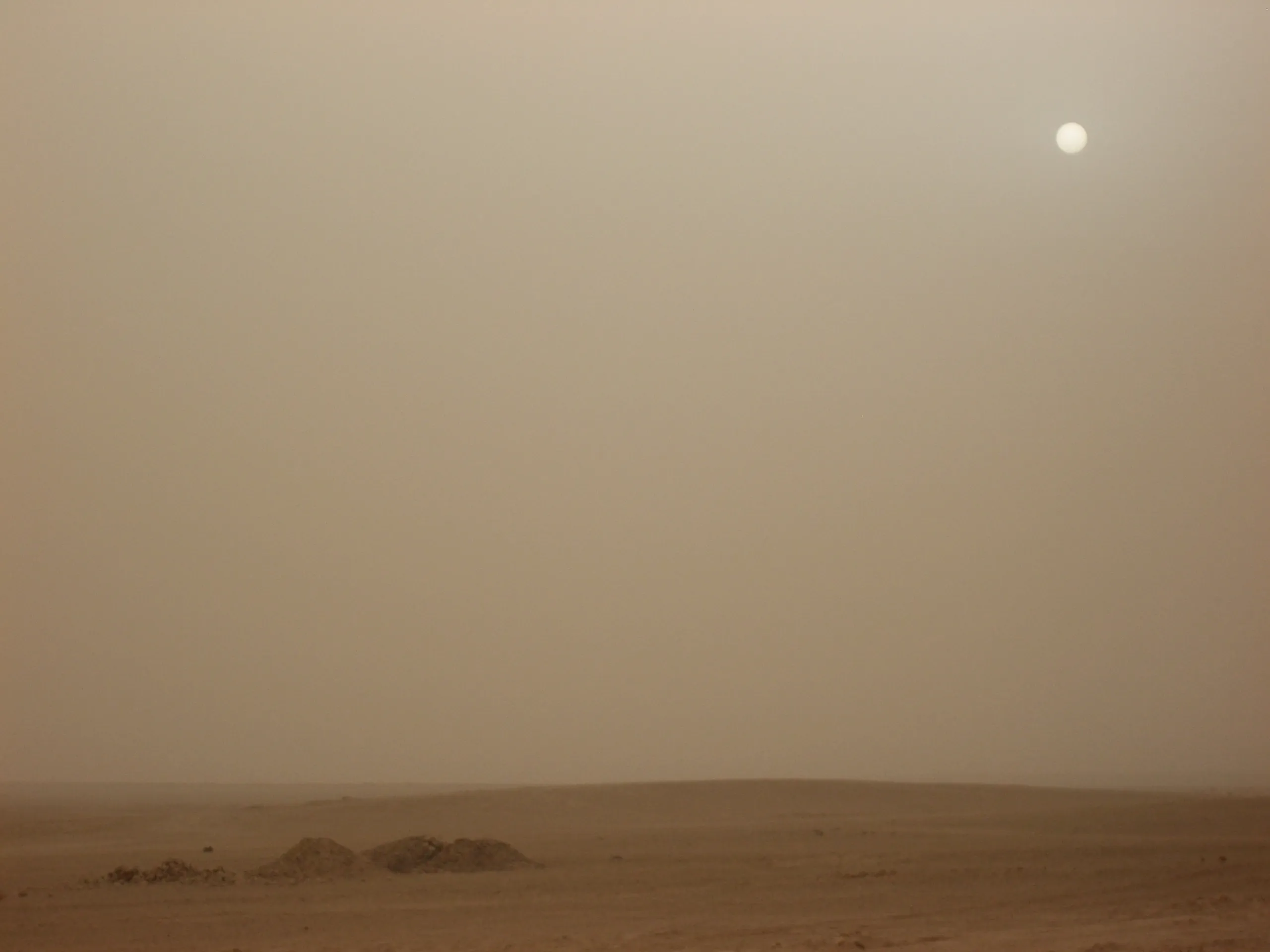 A pale sun shines through dusty haze above a flat desert plain with low mounds near the horizon.