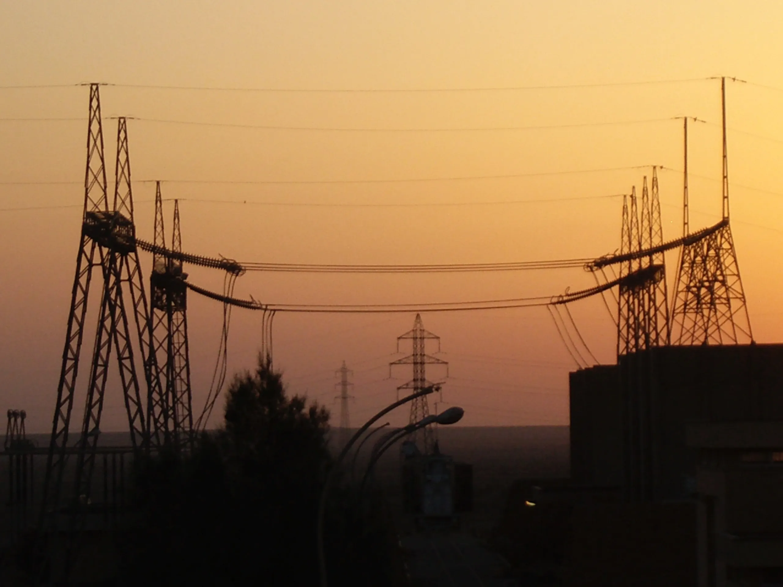 Utility towers, insulators, and suspended power lines silhouetted against an orange dusk sky.