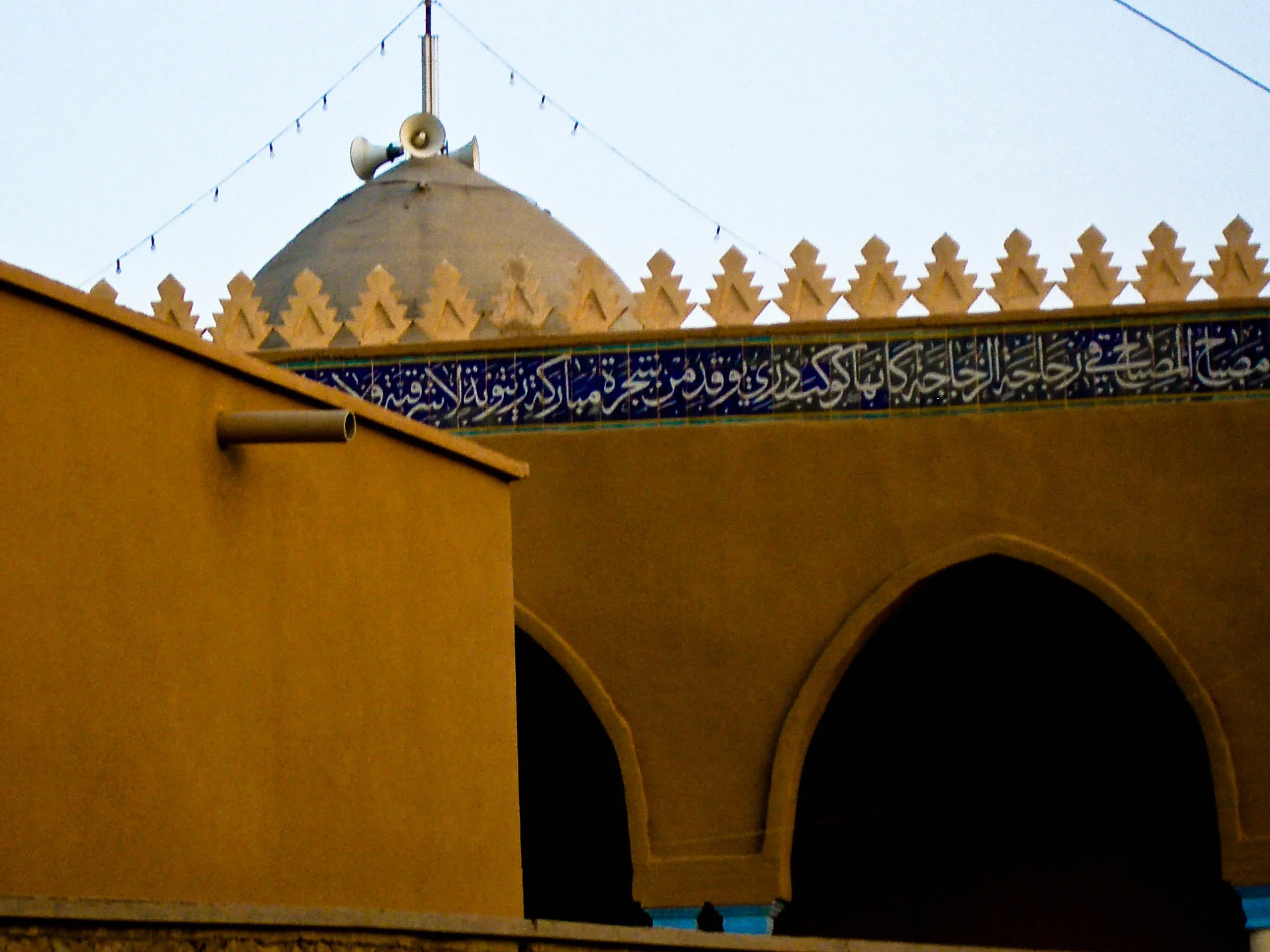 A pale dome rises behind ochre walls with arched openings and a blue band of decorative Arabic calligraphy.