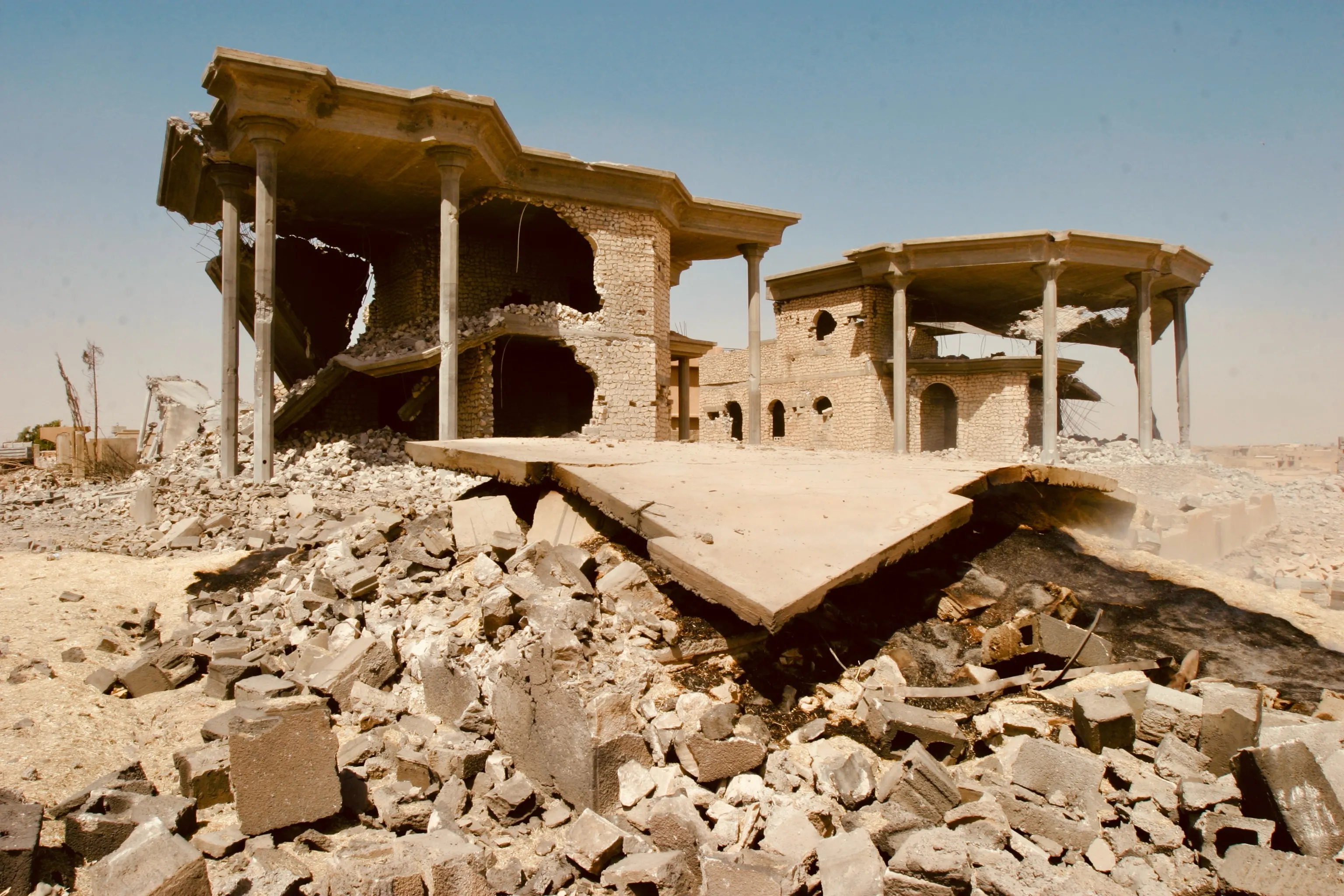 A severely damaged two-story building with broken walls and piles of rubble in a dry landscape.
