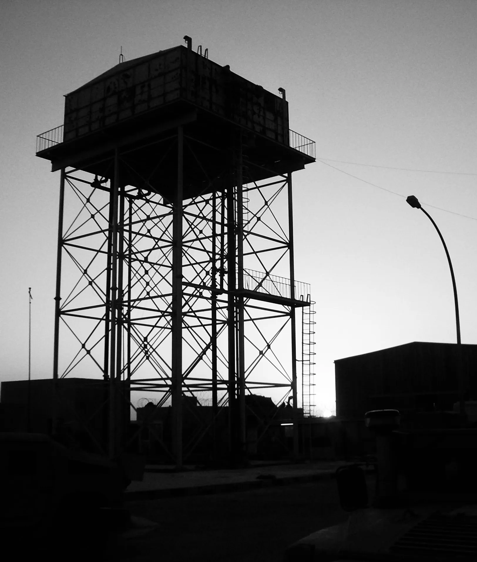 Black-and-white silhouette of a tall industrial water tower beside a curved streetlight at dusk.