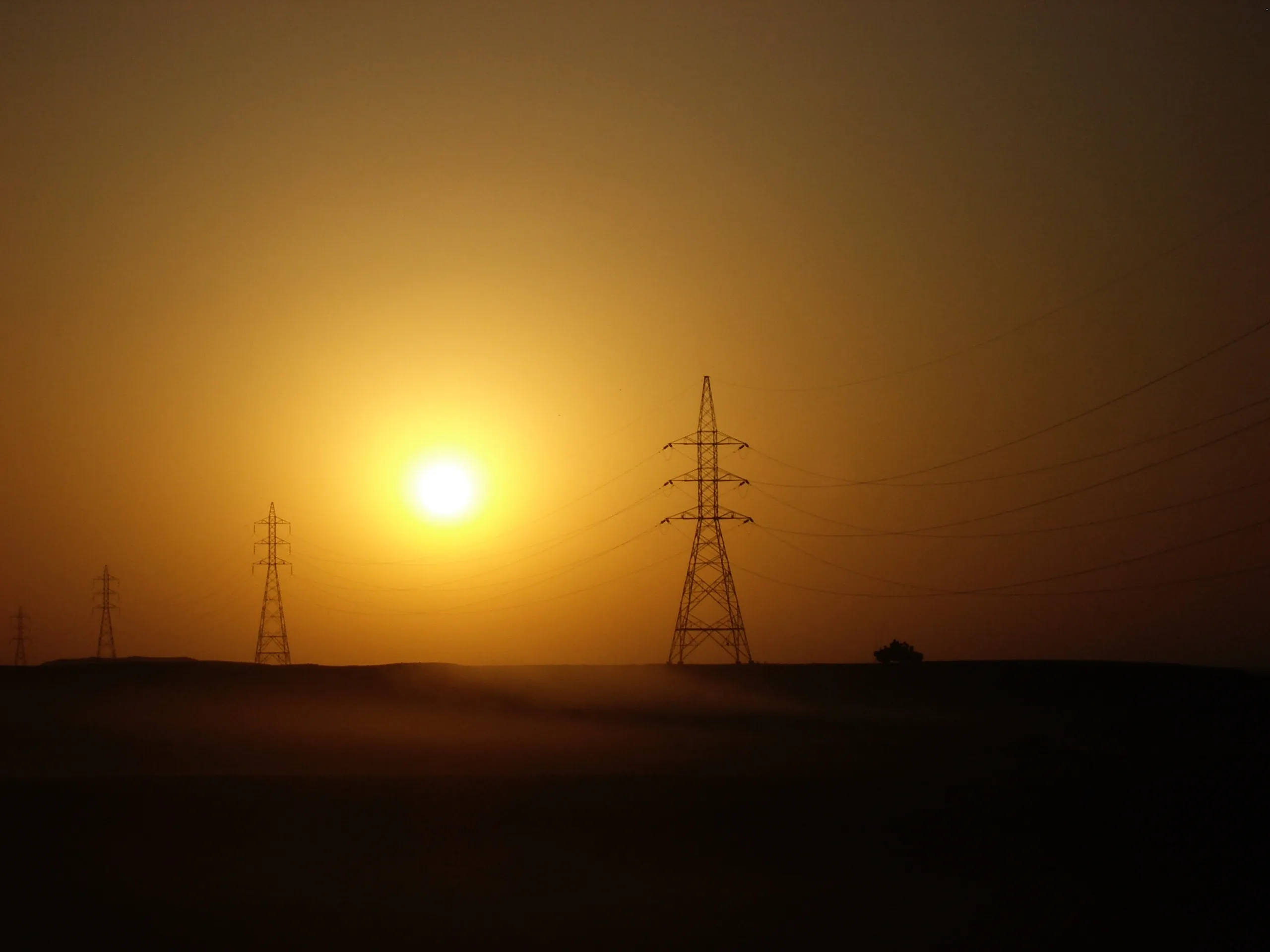 The sun hangs low in an orange sky behind several electricity pylons and power lines.