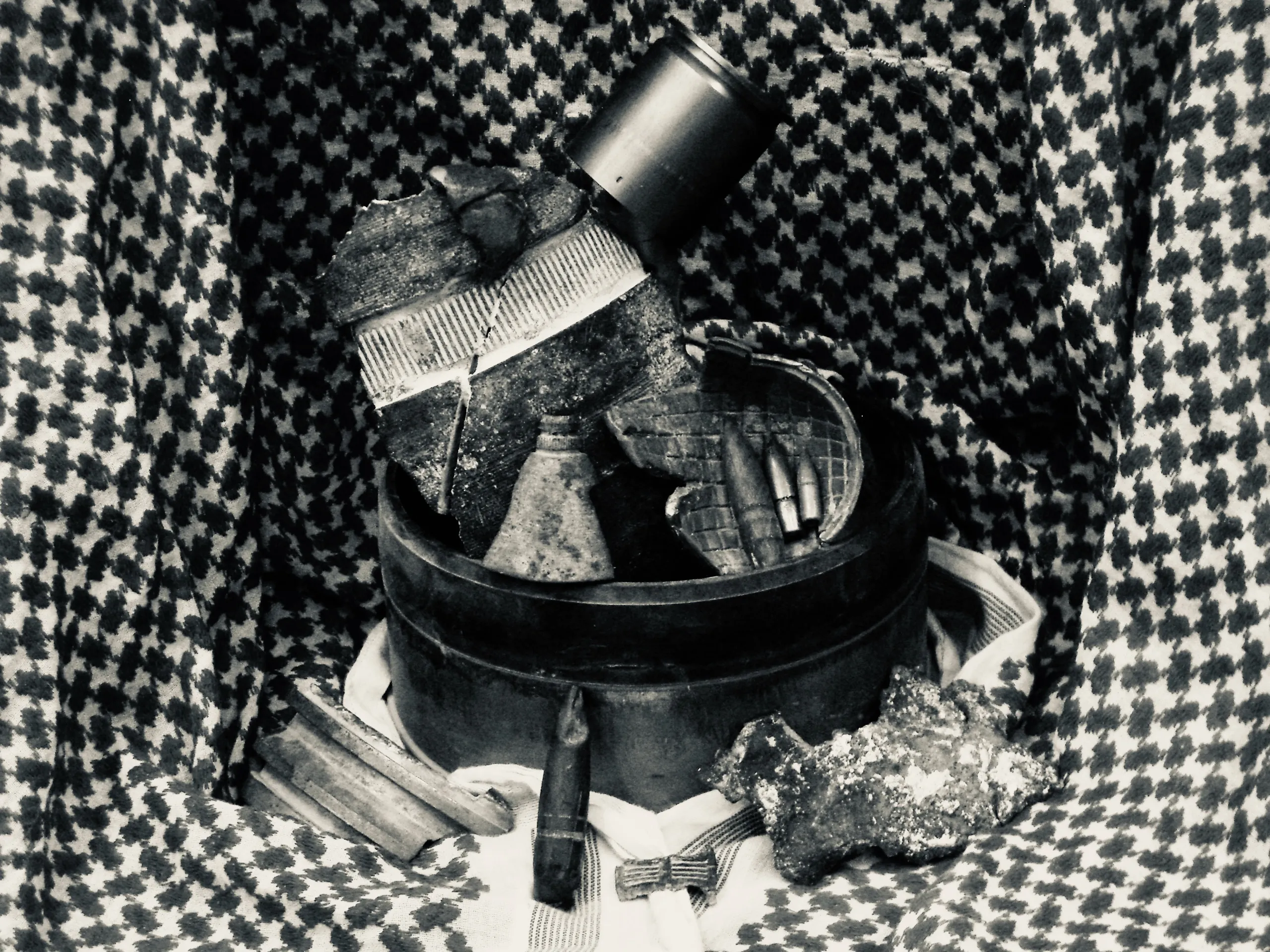 Black-and-white close view of a round case holding small objects and ammunition on patterned cloth.