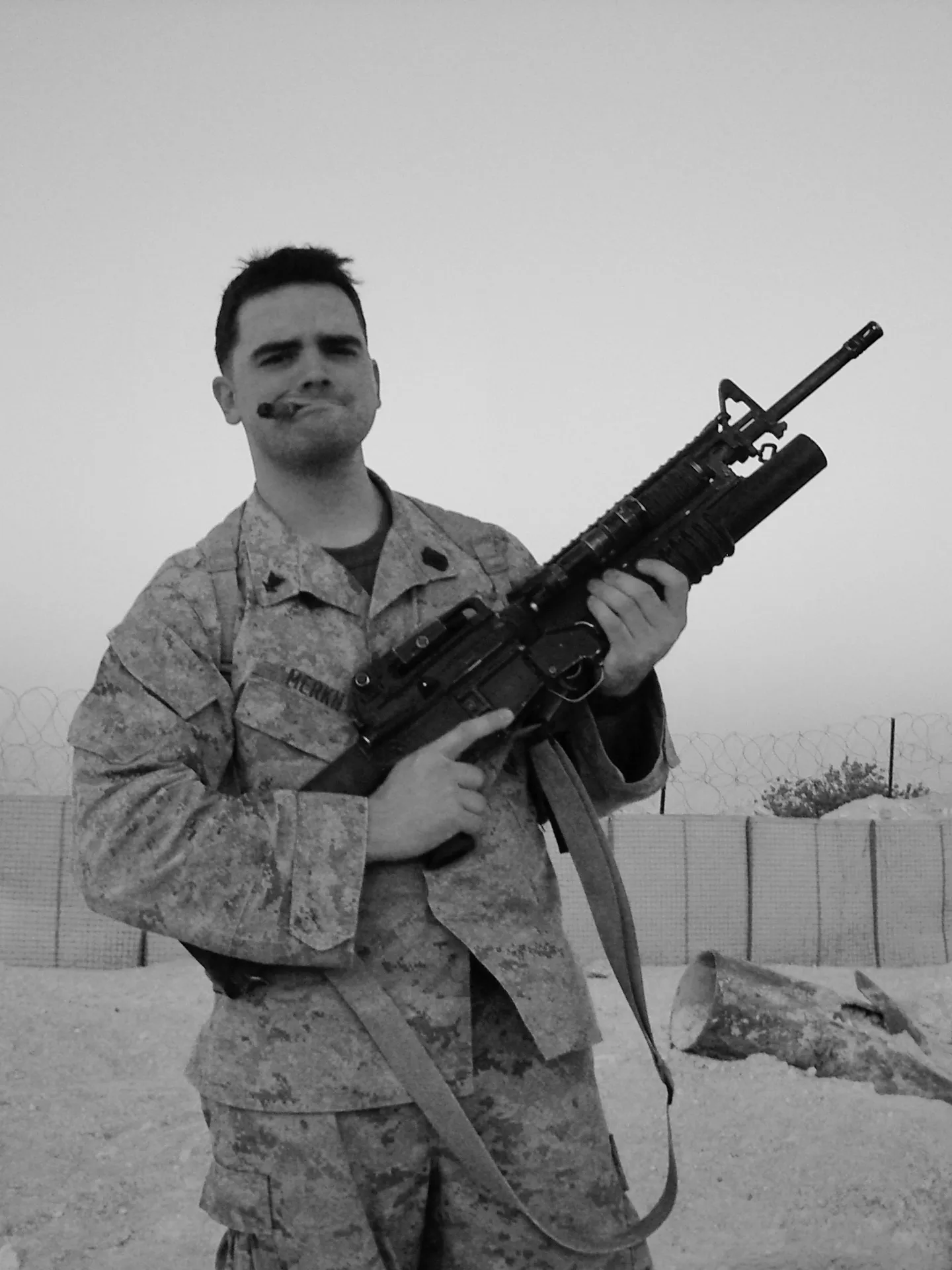 A person in military uniform stands outdoors holding a rifle near barriers and a wire fence.