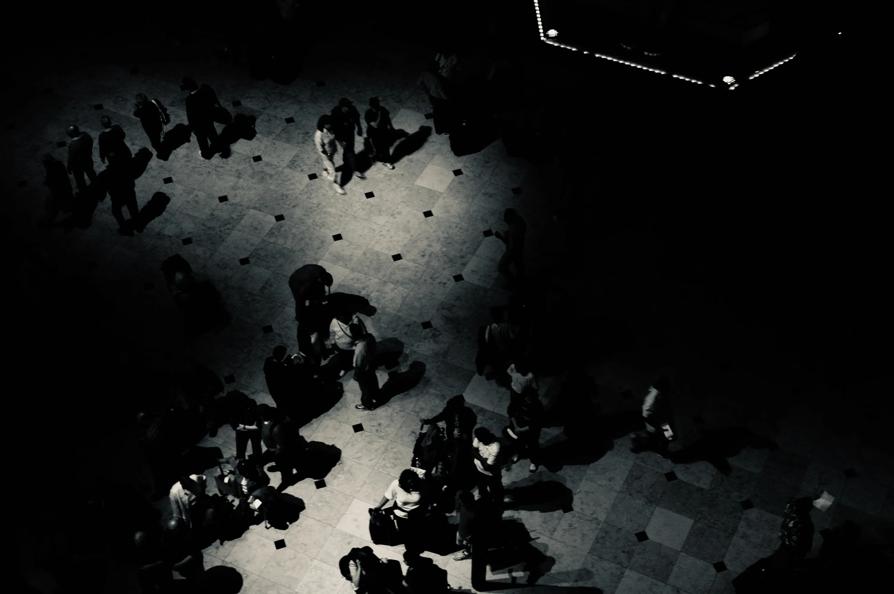 High black-and-white view of people standing and sitting in pools of light on a tiled floor, surrounded by deep shadow.