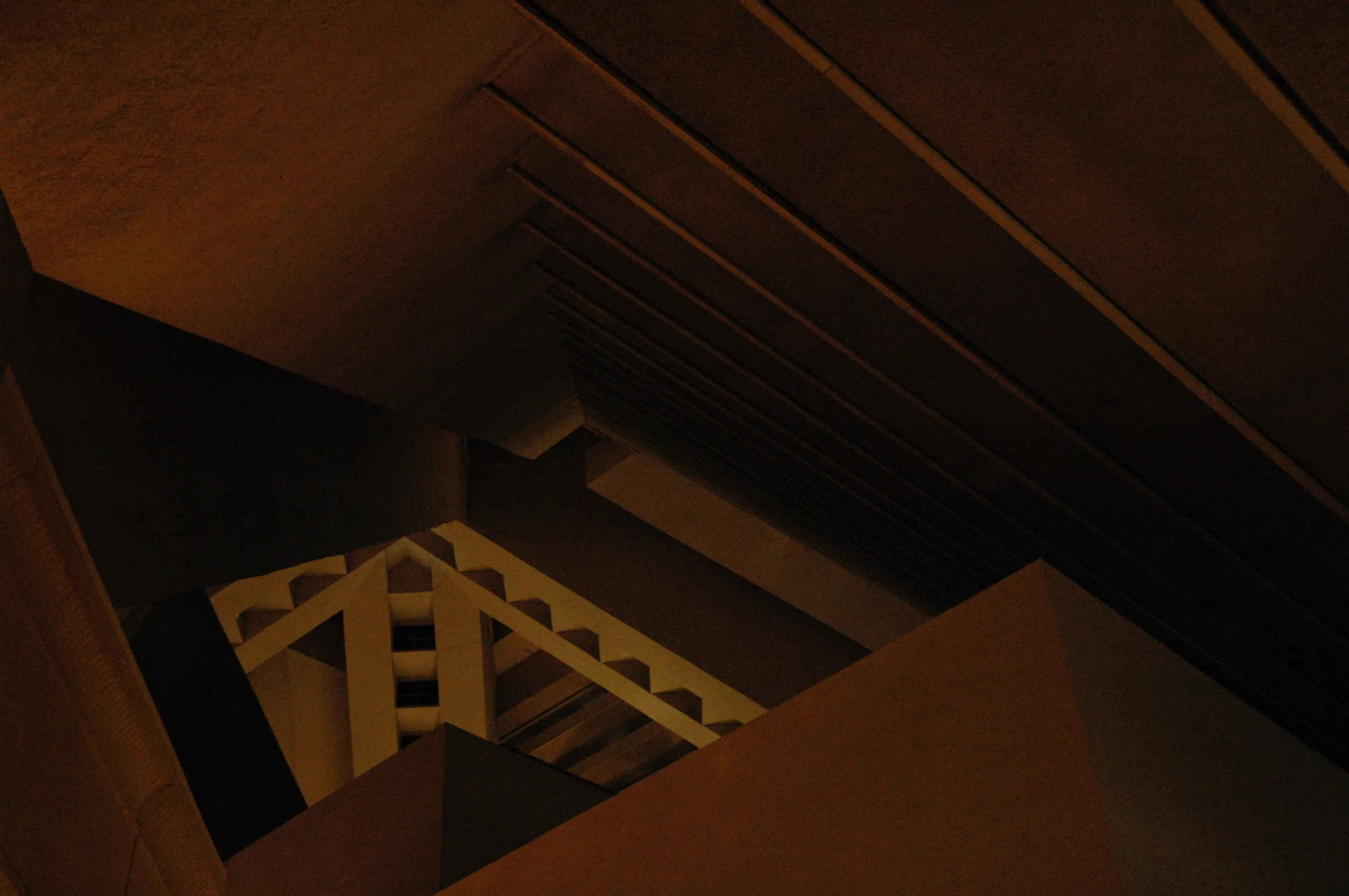 Dimly lit interior view looking down through angular stair rails, beams, and shadowed walls.