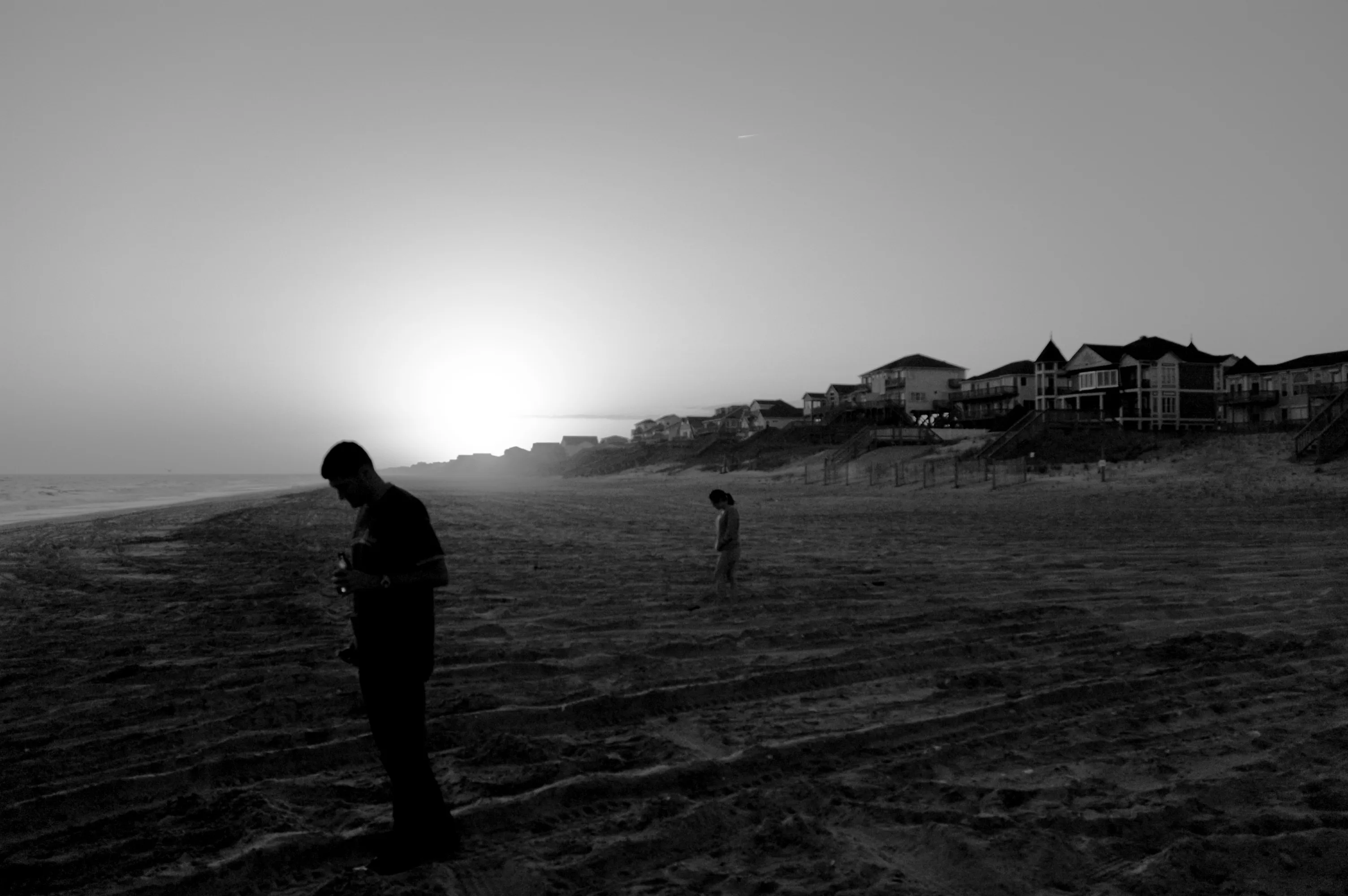 Two people stand apart on a beach near houses at sunset in a black-and-white photograph.