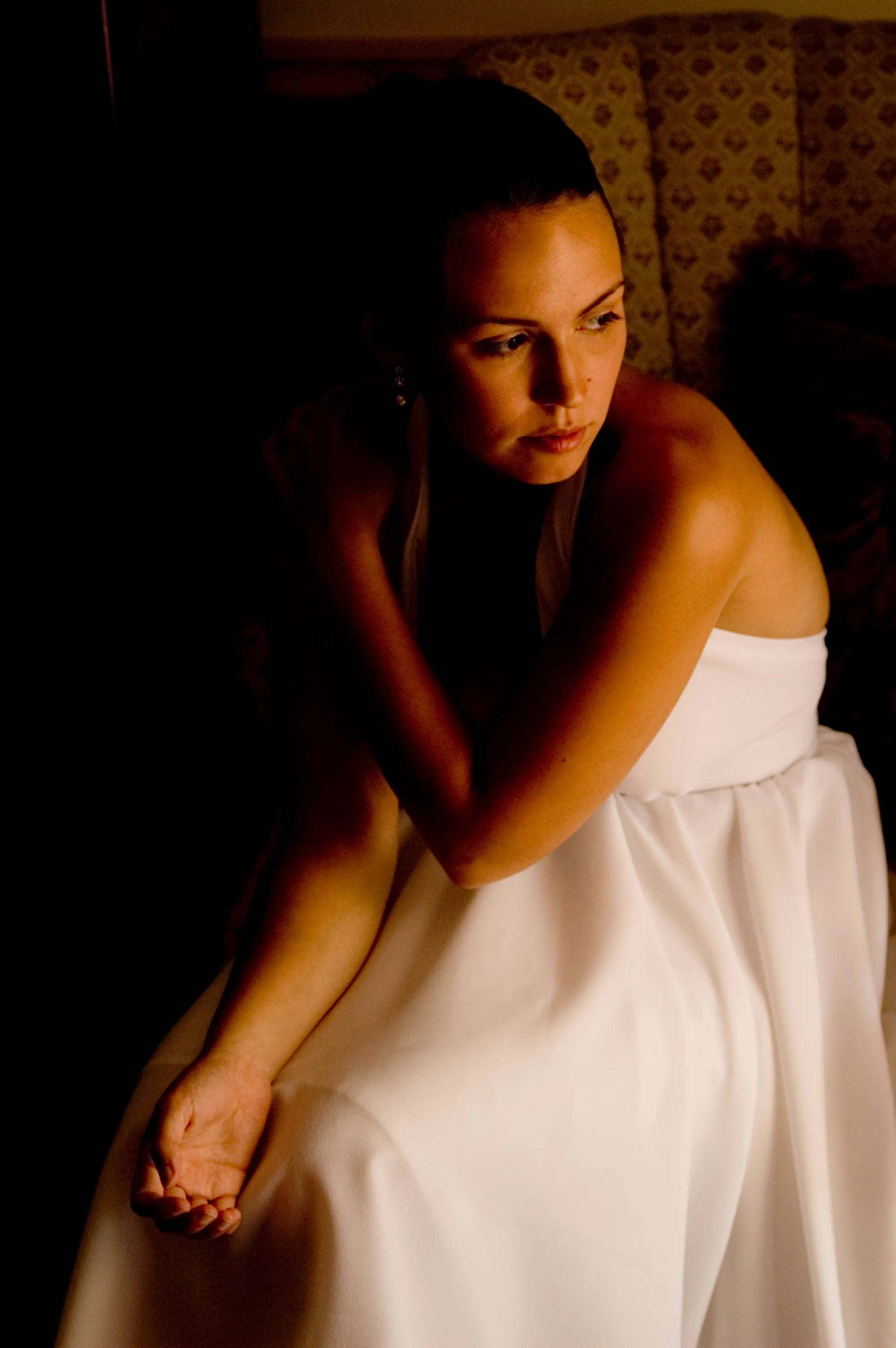 A person in a white dress sits in a dim room, lit warmly from the right and looking off to the side.