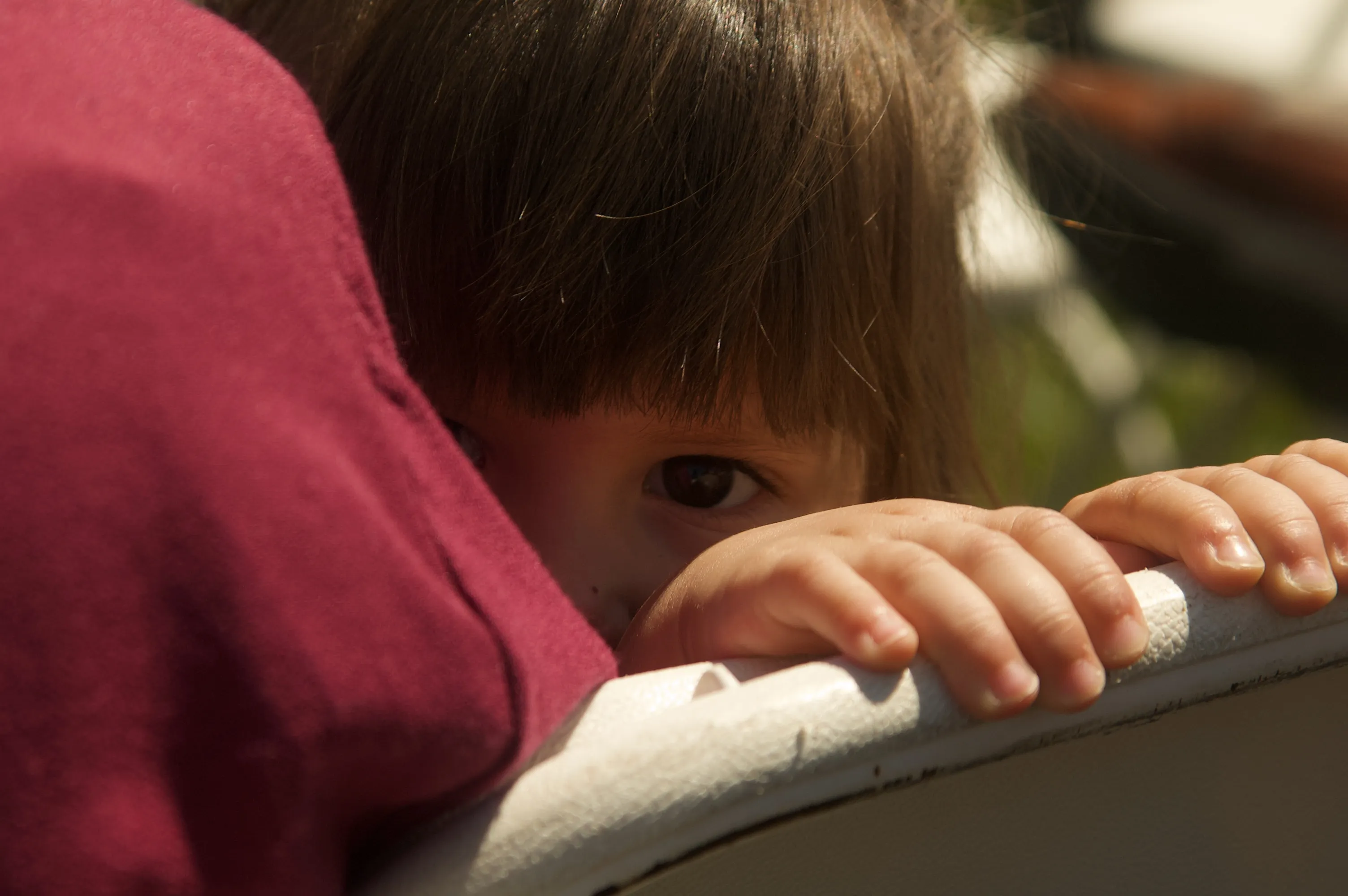 A young child peering over a white railing, with hands on the rail and one eye visible behind an adult’s shoulder.