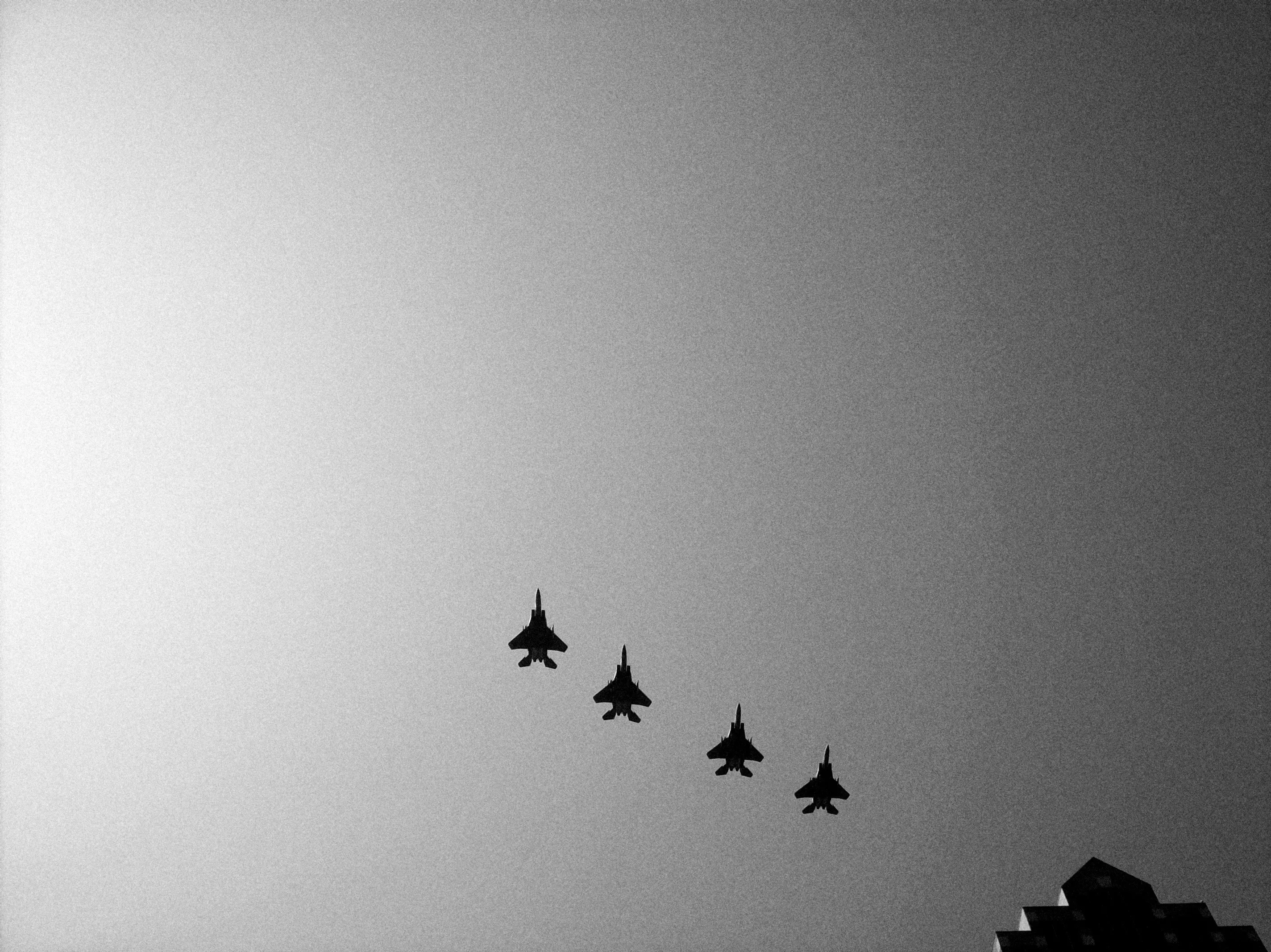 Four dark fighter jets in formation against a pale gray sky, with the corner of a building at lower right.