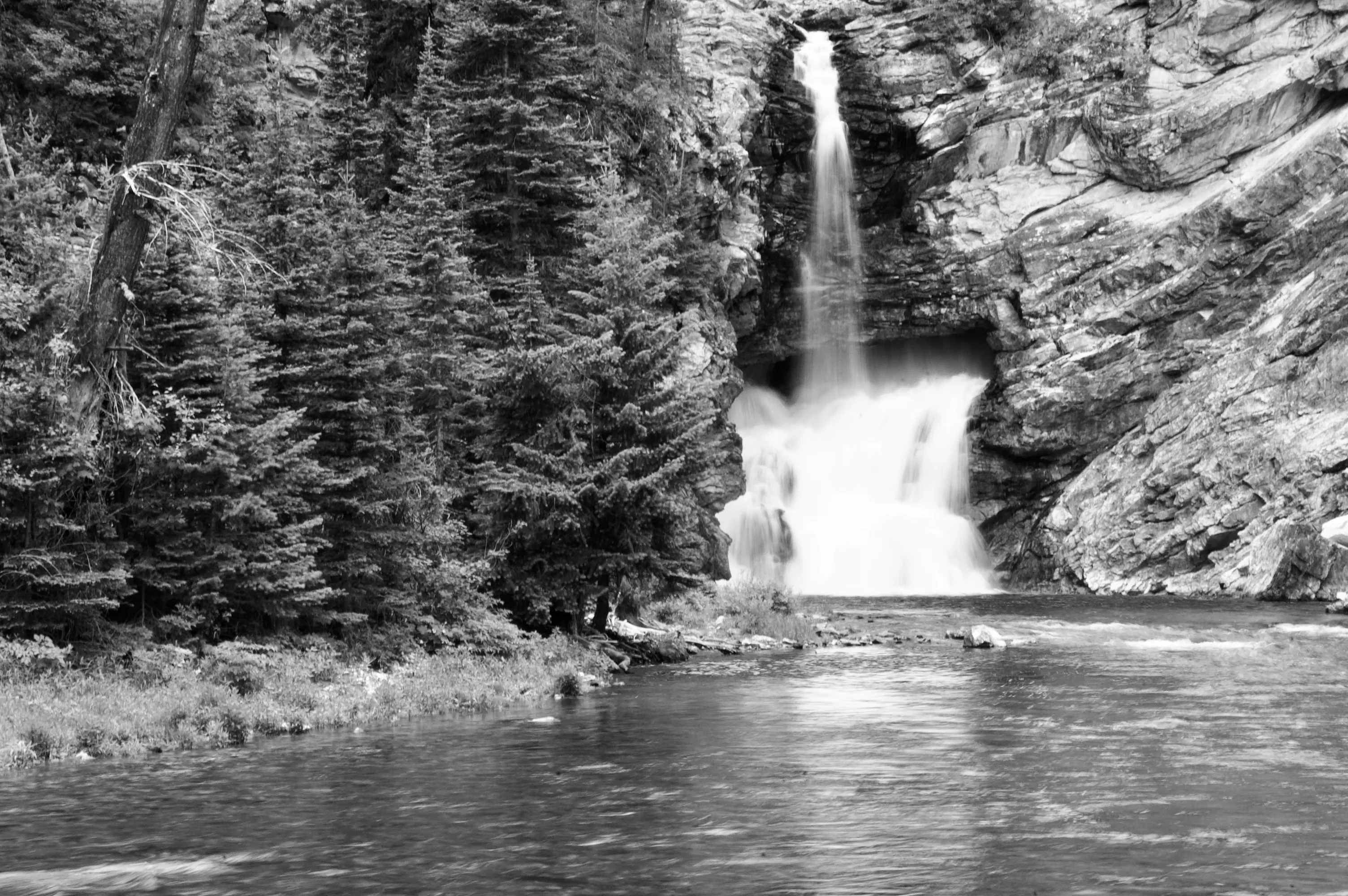 Black-and-white photograph of a waterfall pouring into a river beside dense conifers and steep rock walls.