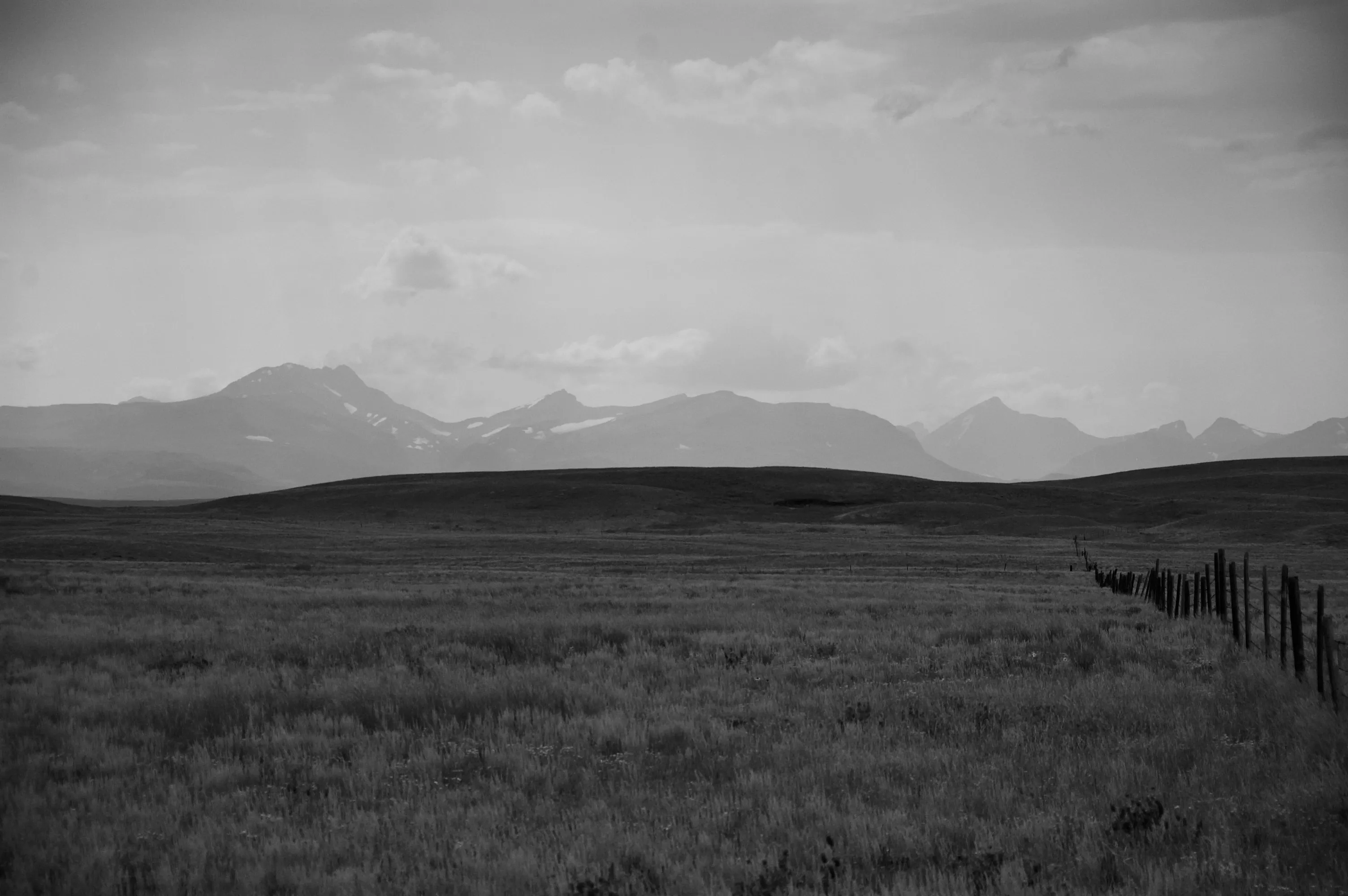 Black-and-white view of a grassy plain with a fence line leading toward distant mountains under clouds.
