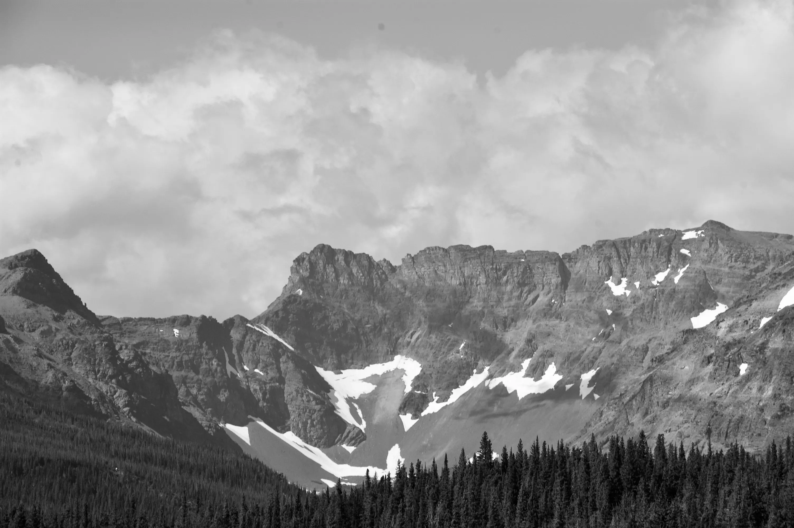 Black-and-white landscape of a rugged mountain ridge with snow patches above a dark conifer forest.
