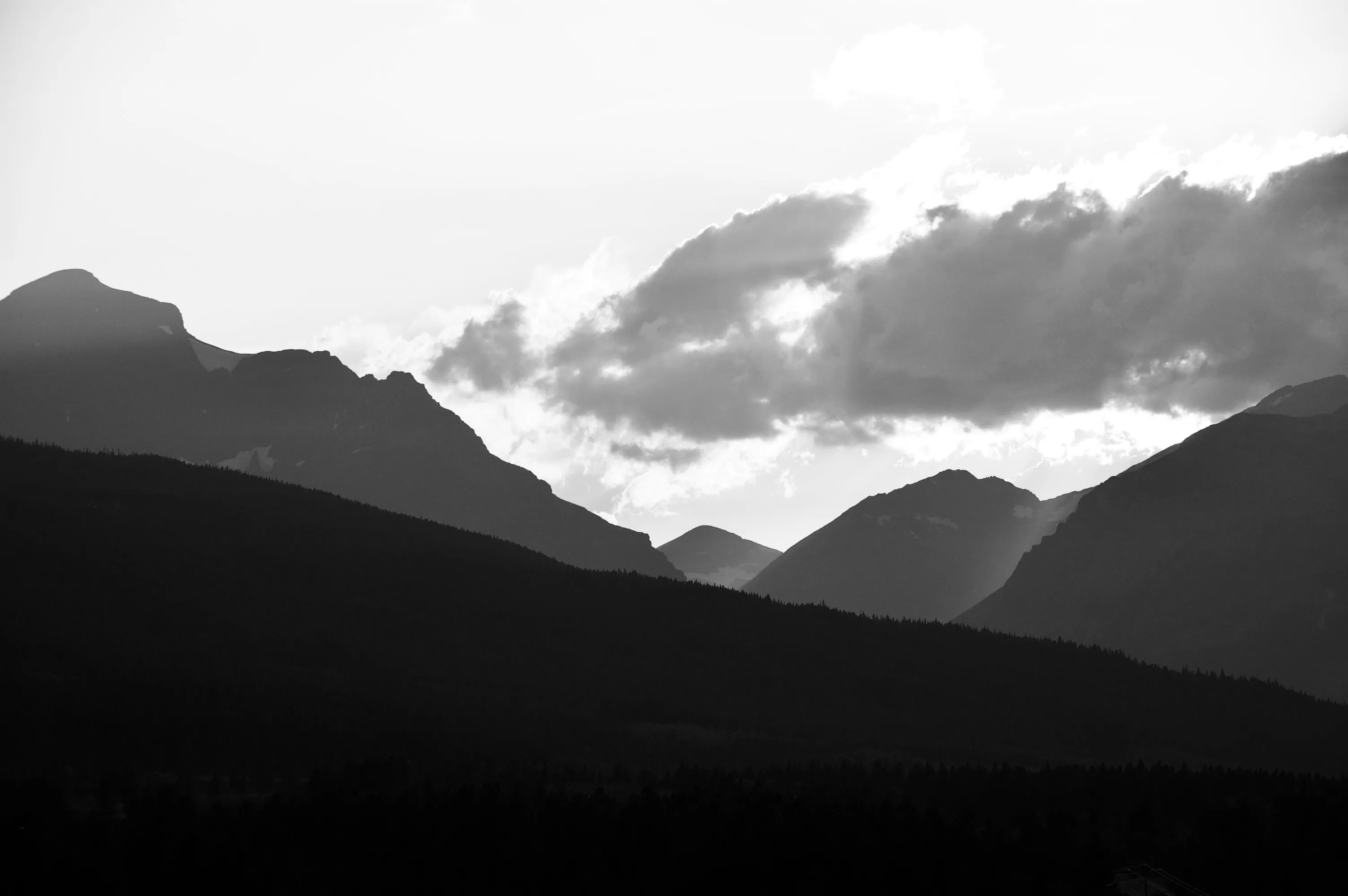 Black-and-white landscape of overlapping dark mountain ridges below bright clouds.