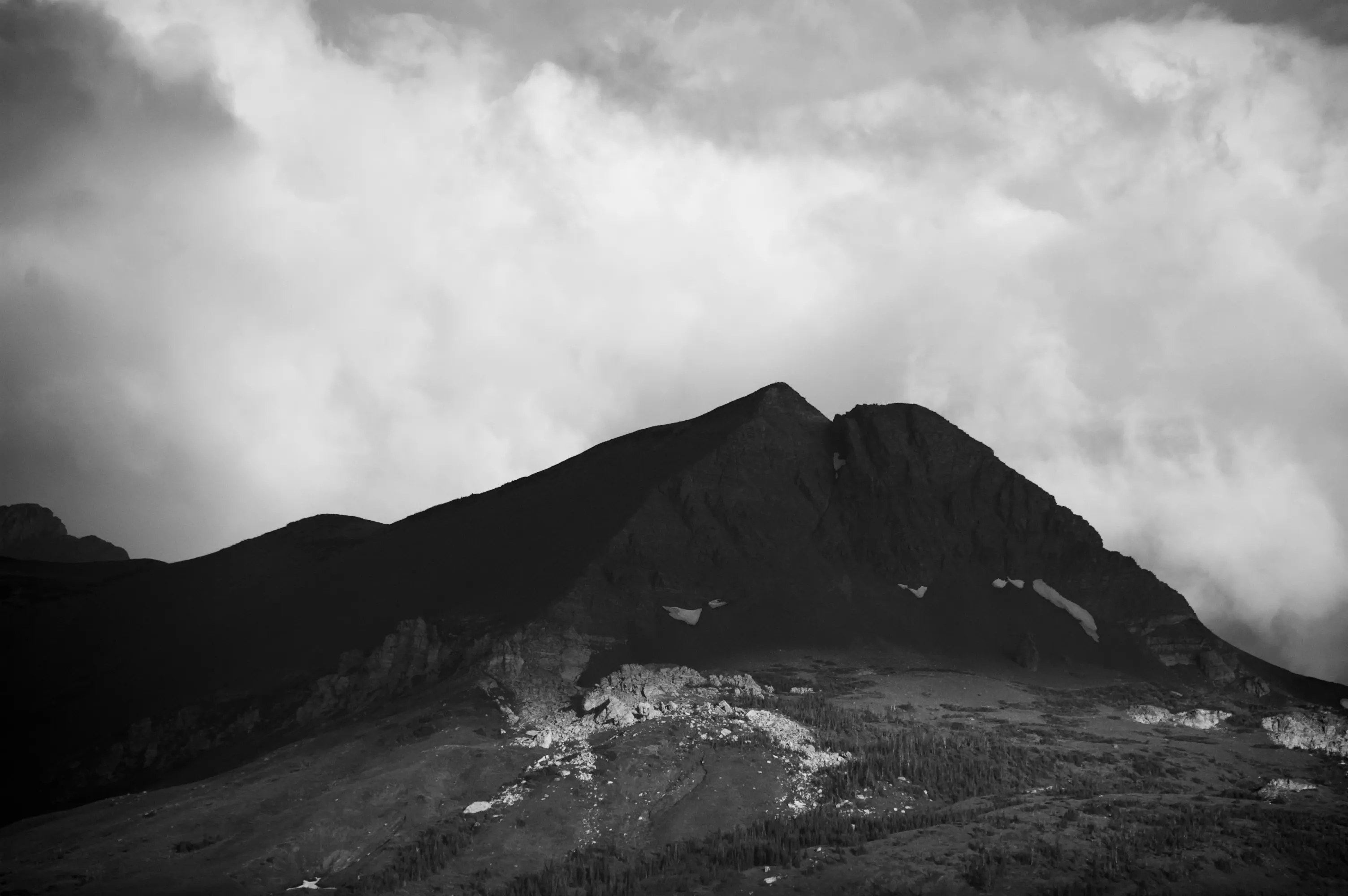 Black-and-white photograph of a steep dark mountain ridge beneath thick, luminous storm clouds.