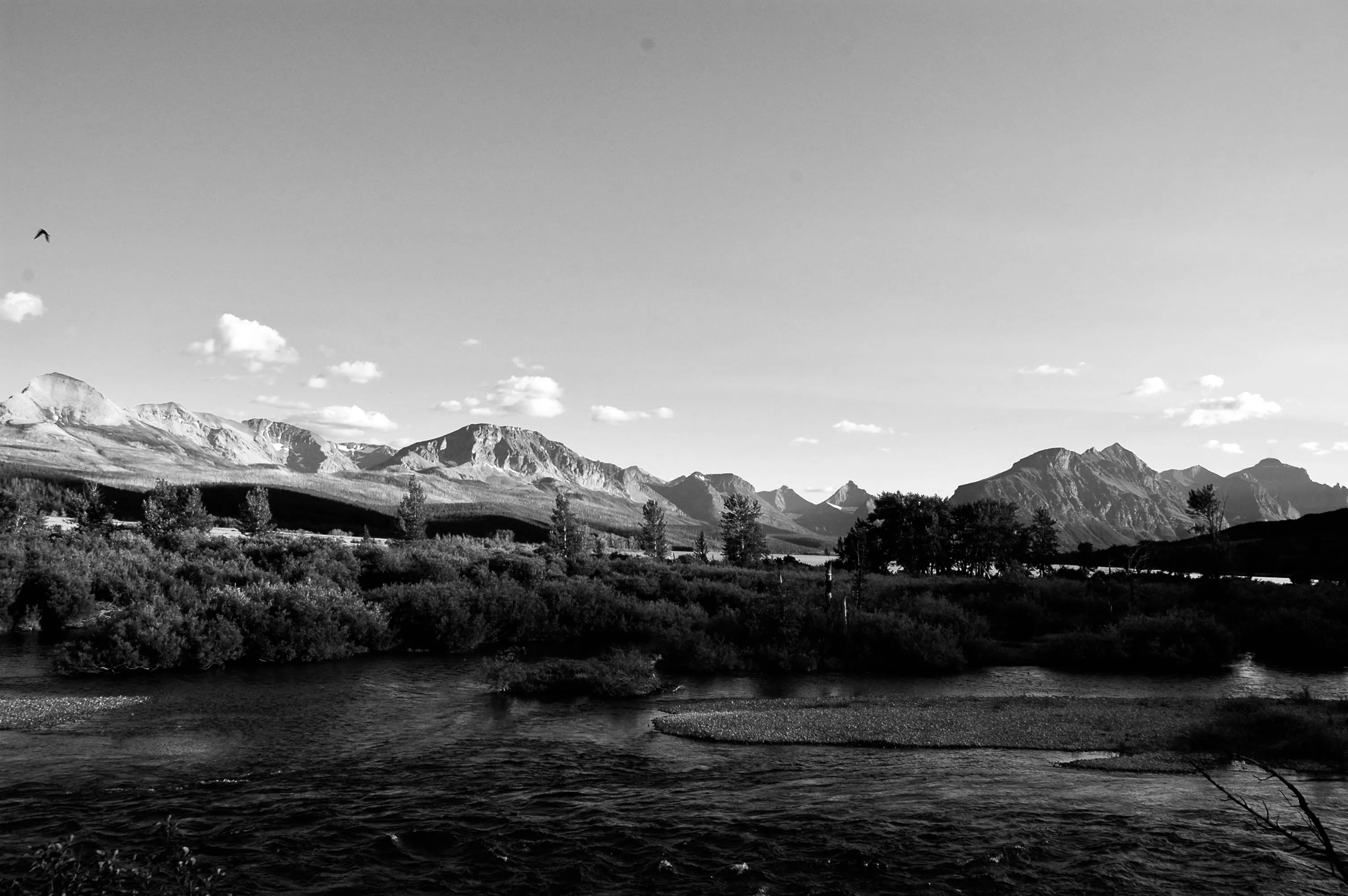 Black-and-white landscape of a river, low trees, and jagged mountains beneath scattered clouds.