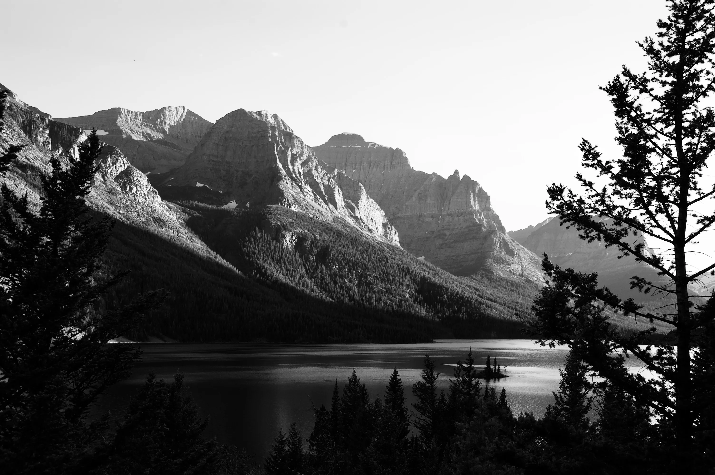 Black-and-white landscape of steep mountain peaks above a calm lake, framed by dark conifer trees.