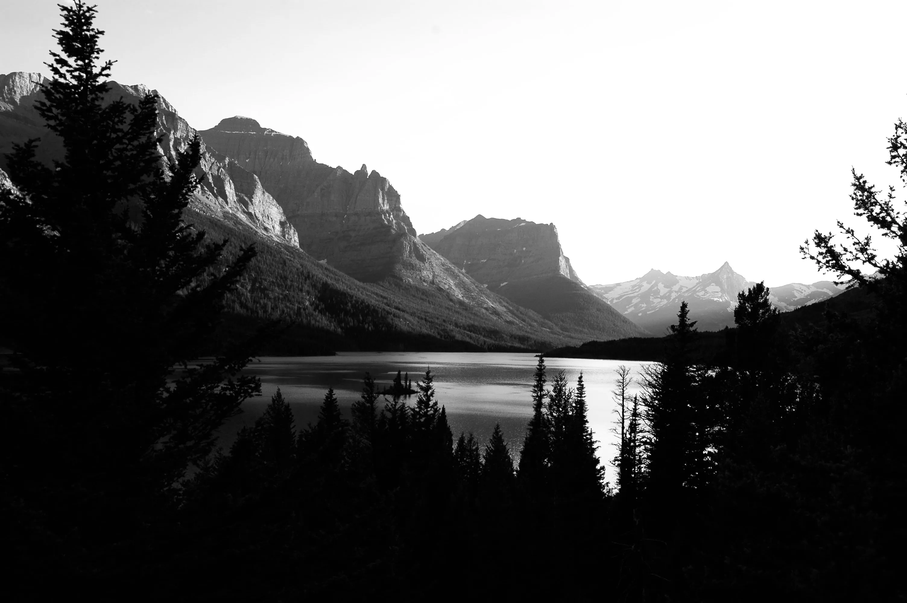 A calm mountain lake sits beneath steep rocky peaks, framed by dark evergreen trees.