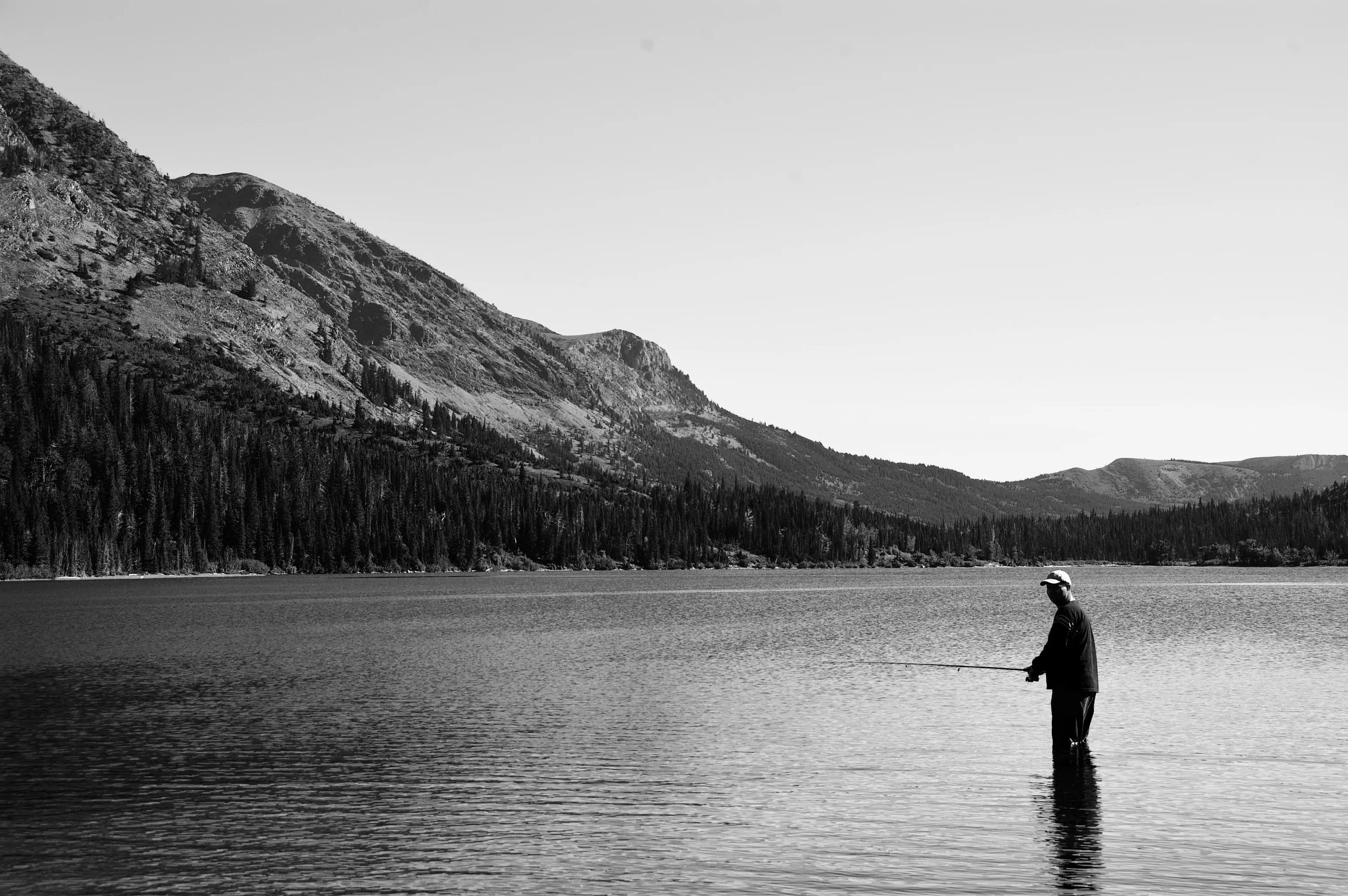 Black-and-white photograph of a person standing in shallow water and fishing in a lake beneath steep forested mountains.