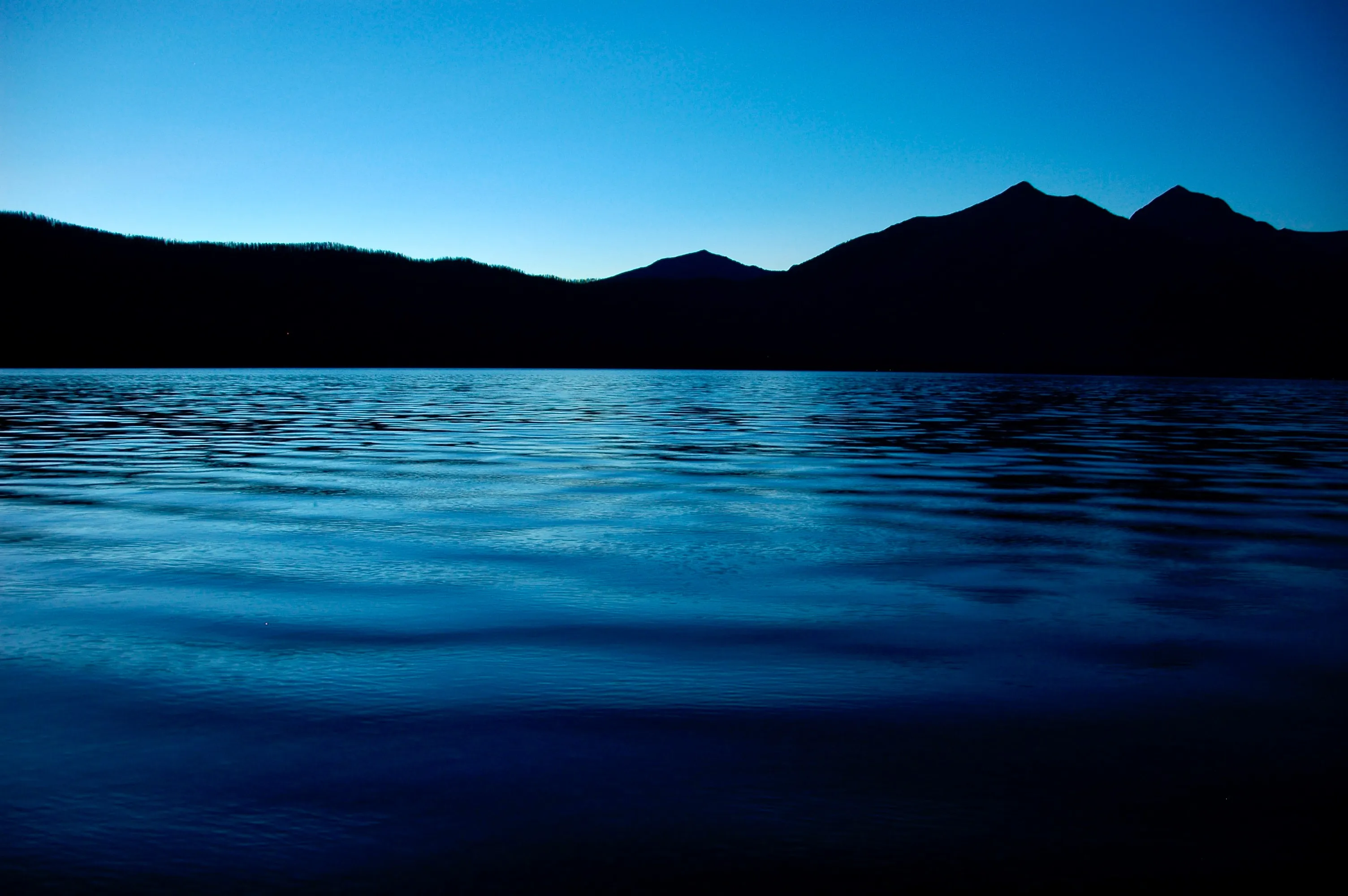 Blue water fills the foreground beneath dark mountain silhouettes and a deep blue evening sky.