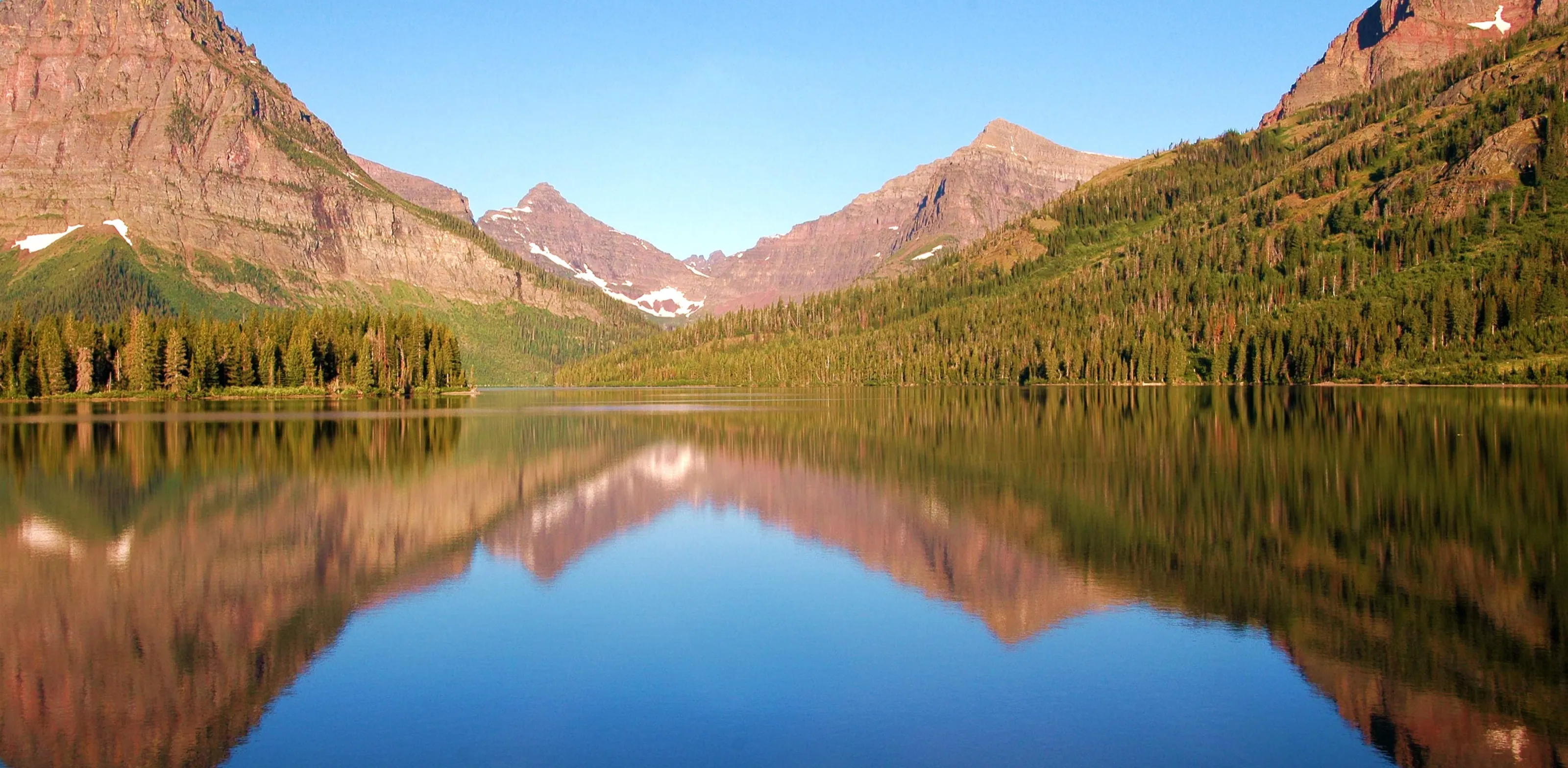 Pink-lit mountain peaks and evergreen forest are reflected almost symmetrically in a calm lake.