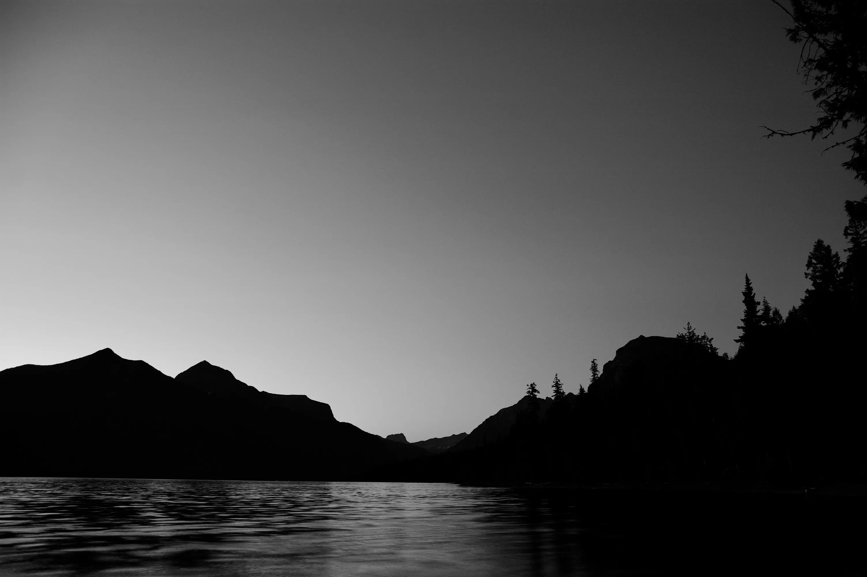 Black-and-white lake at dusk with dark mountain and tree silhouettes against a pale sky.
