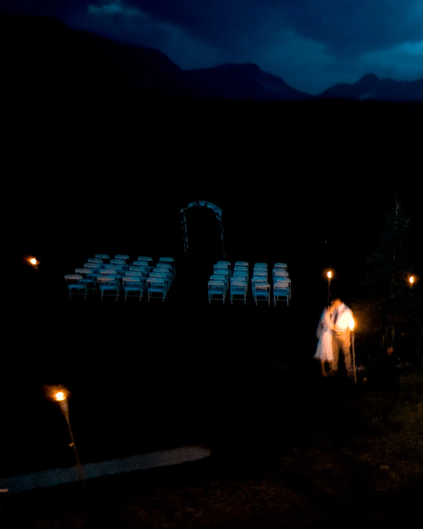 A very dark outdoor scene with rows of chairs, small torches, and a blurred person in light clothing.
