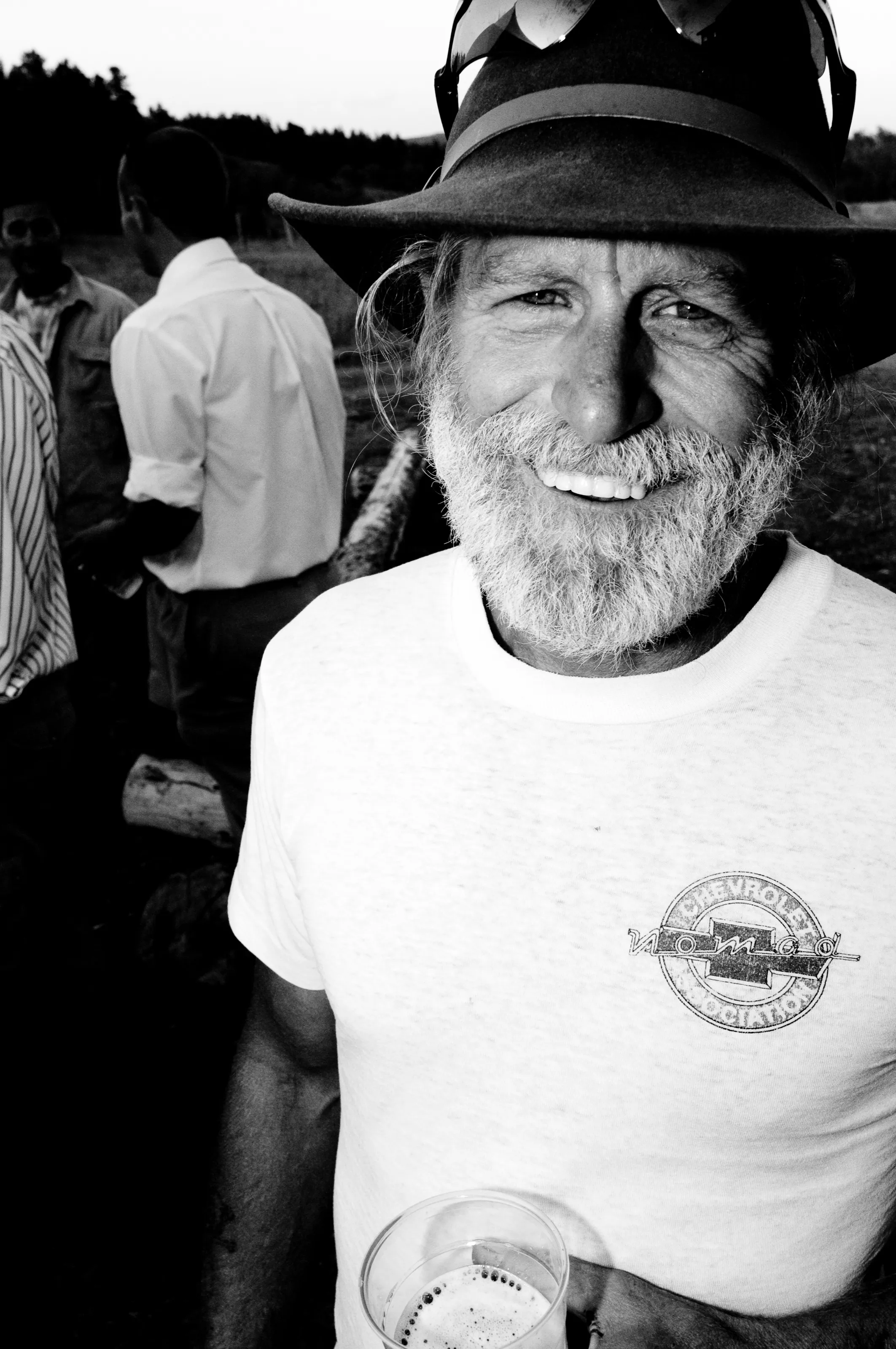 Black-and-white close portrait of a smiling bearded man in a wide-brimmed hat, with other people behind him.