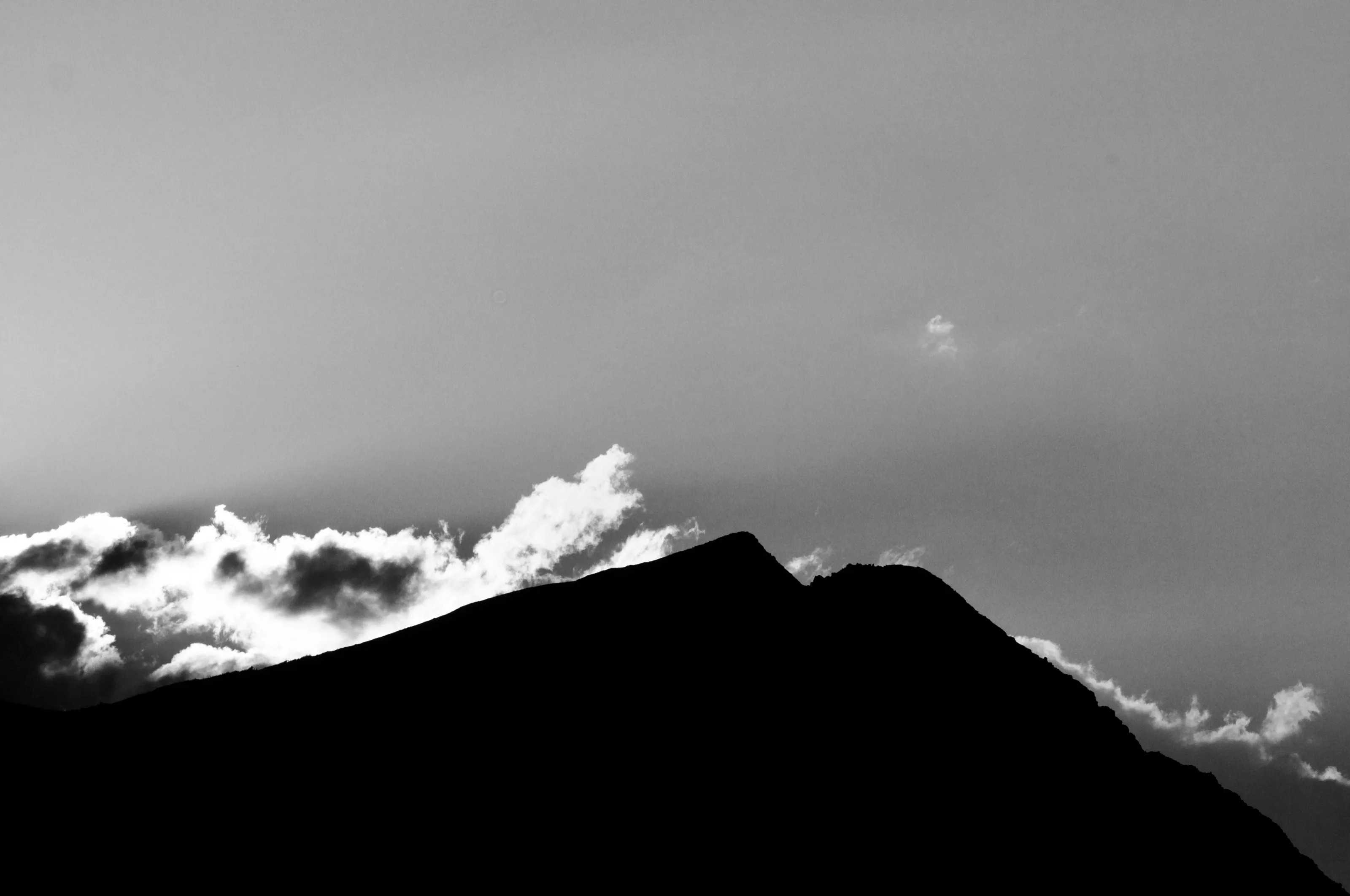 Black-and-white photograph of a dark mountain silhouette against bright clouds and a pale sky.