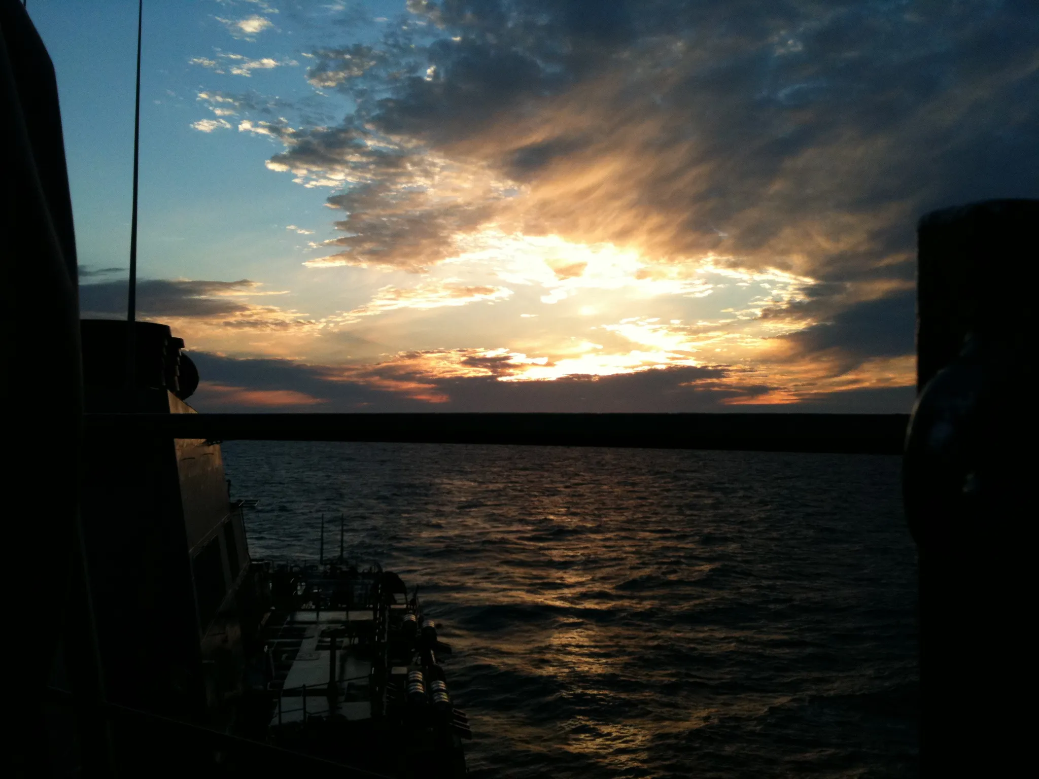 View from a dark ship deck toward a bright sunset over rippled water and low clouds.