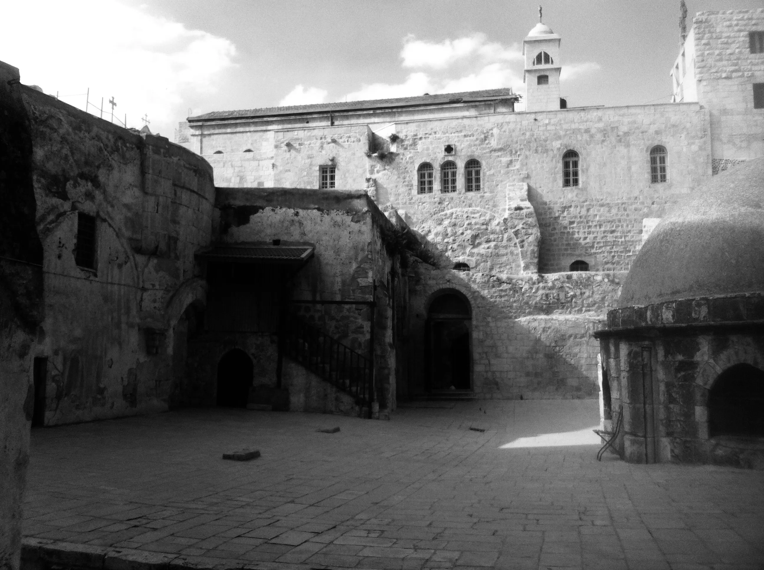 Black-and-white photograph of a paved stone courtyard surrounded by weathered stone buildings and arched doorways.