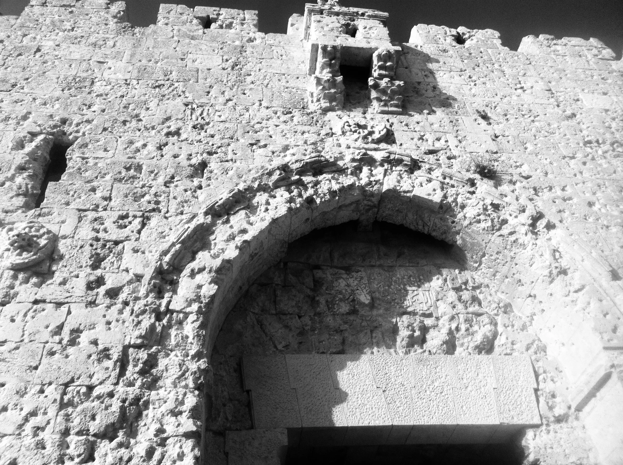 Black-and-white upward view of a crumbling stone wall with a large arched opening and a small doorway above it.