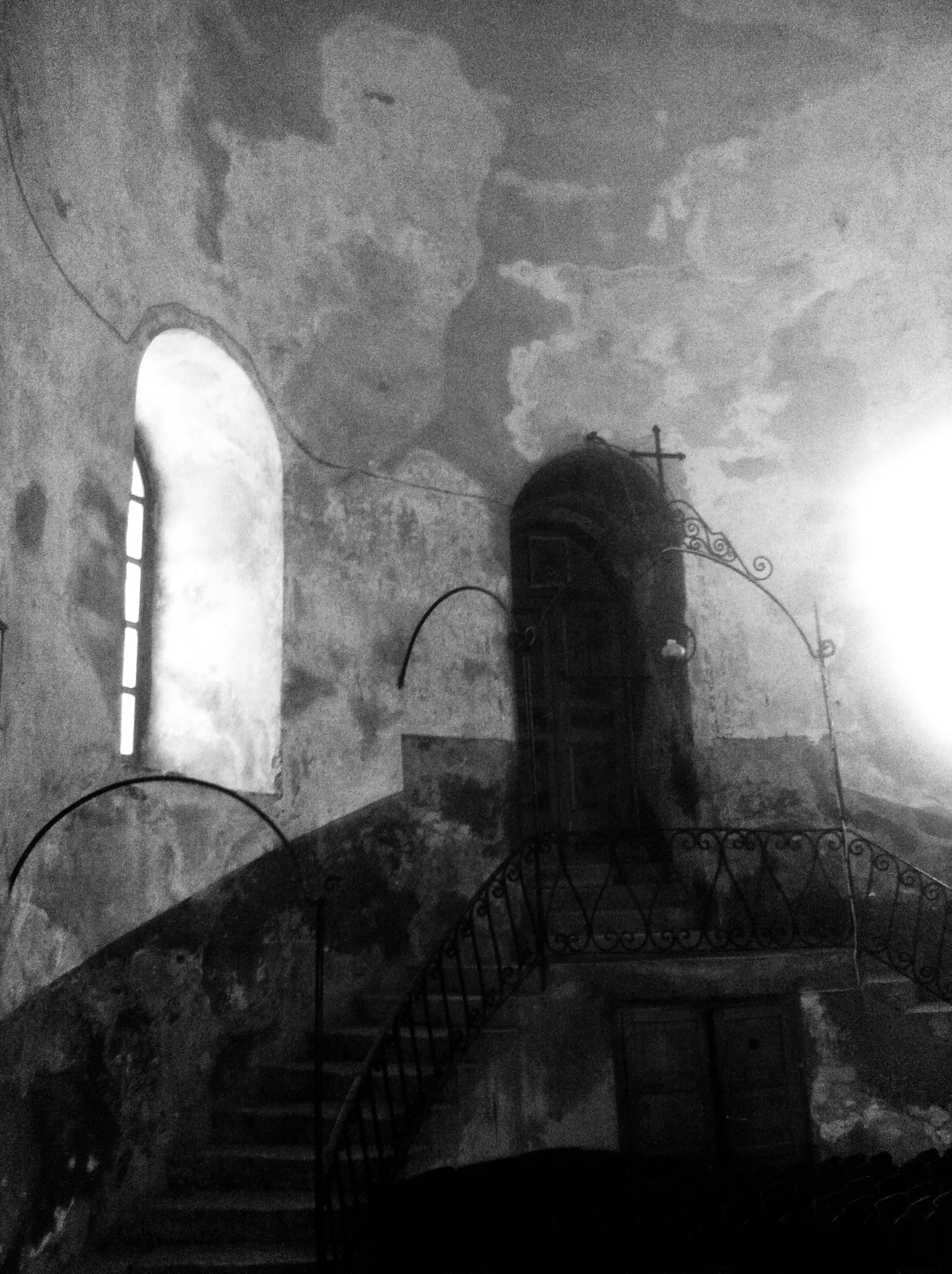 Black-and-white interior of a weathered stone building with a staircase, arched window, dark door, and decorative iron railings.