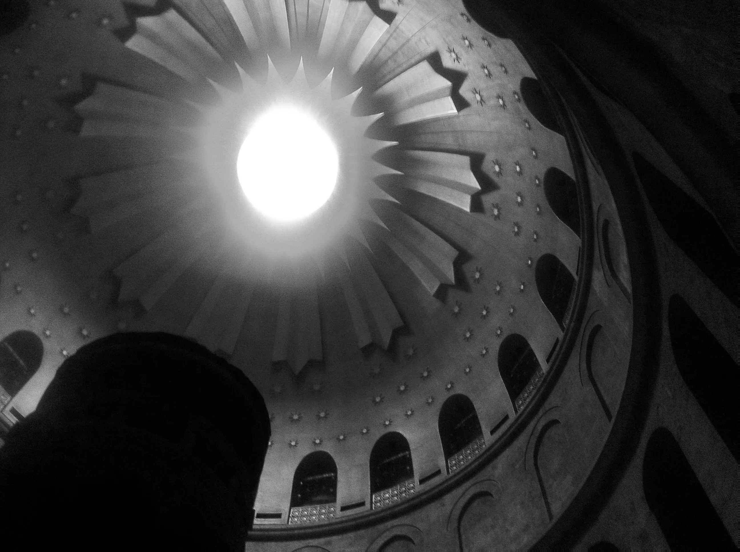 Black-and-white upward view inside a domed building, with a bright circular opening surrounded by radiating architectural forms.