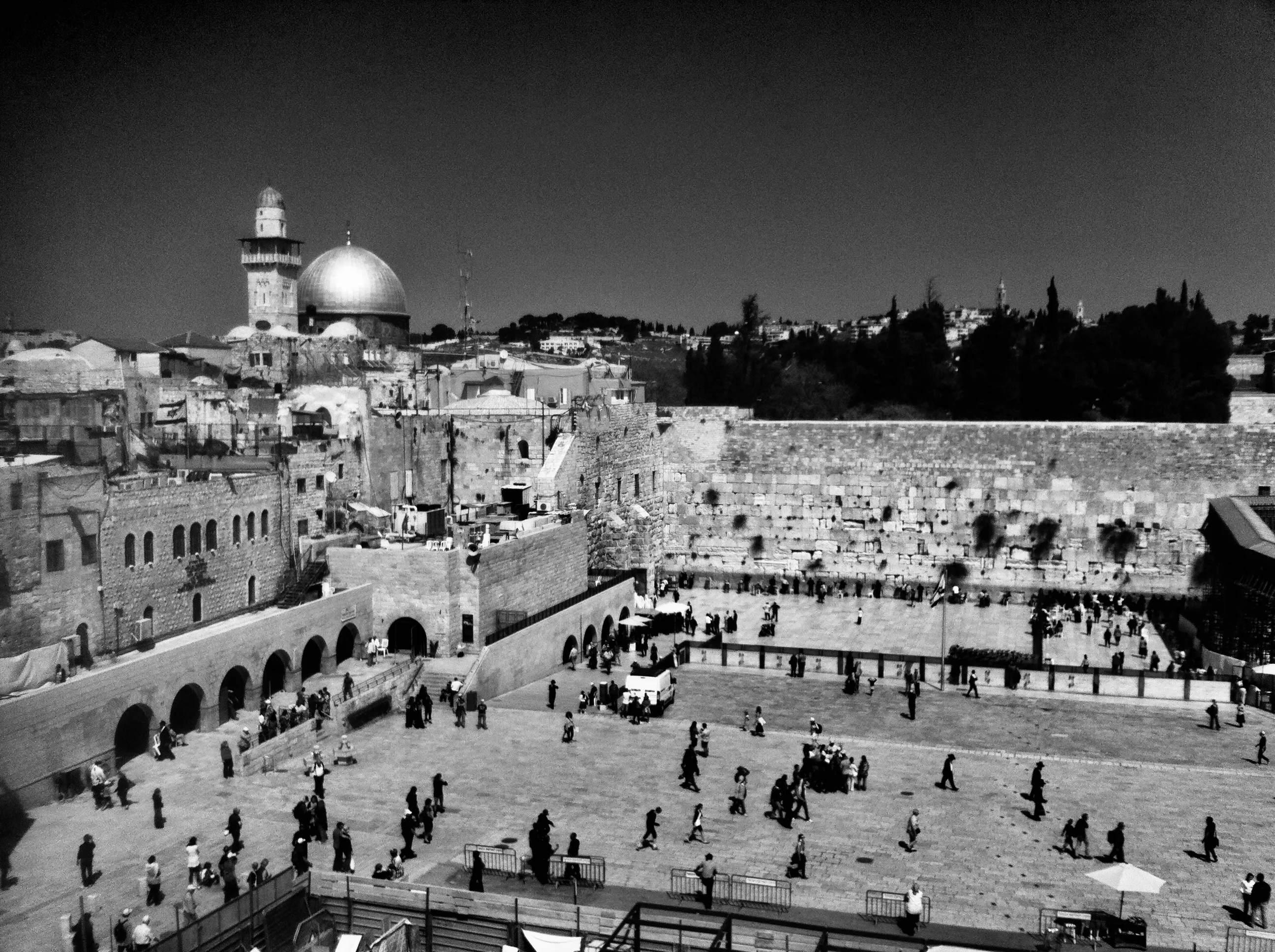 Black-and-white elevated view of many people in a stone plaza beside a large ancient wall, with a dome and tower behind it.