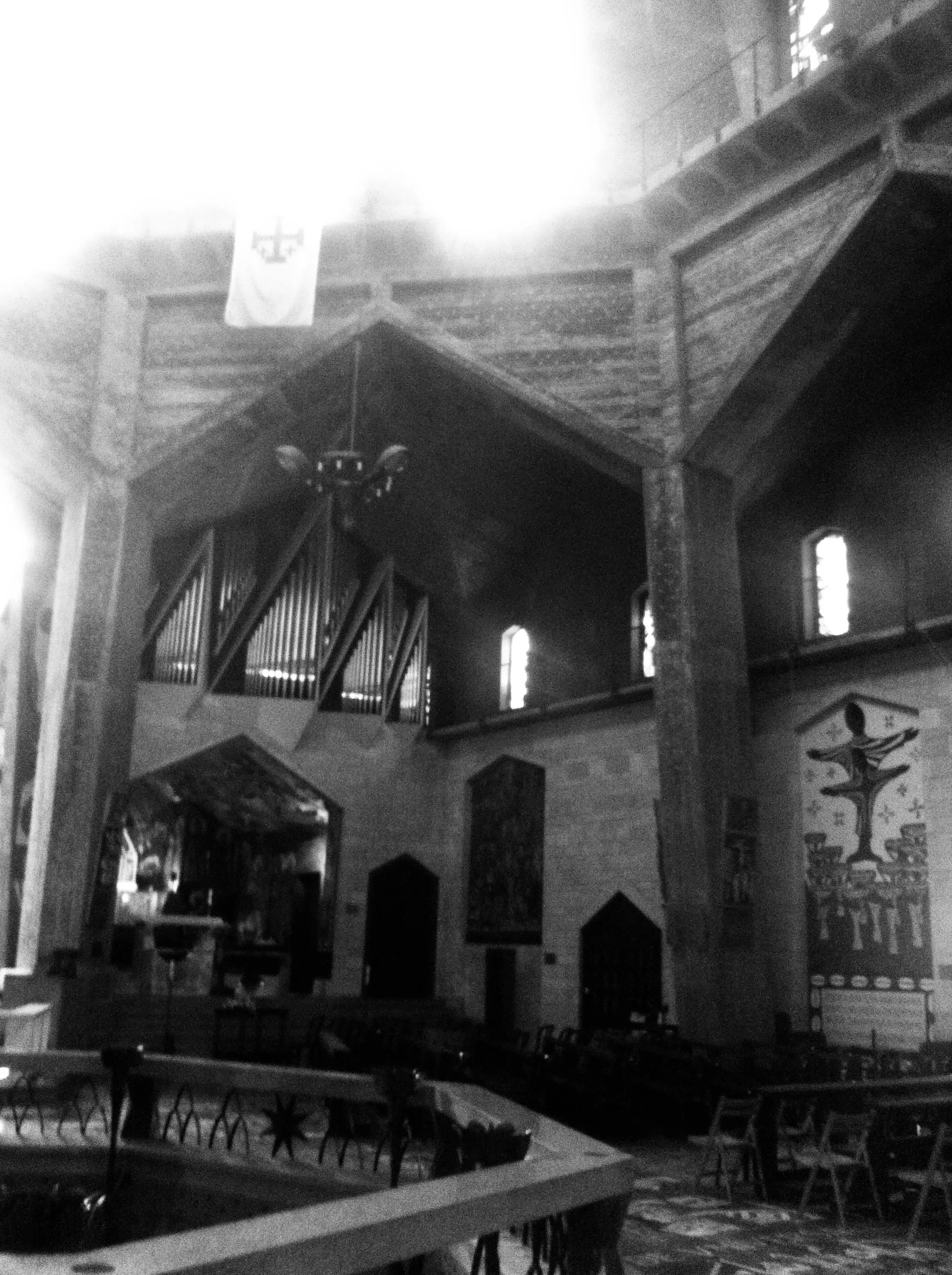 Grainy black-and-white photograph of a church interior with a pipe organ, stone columns, banners, and chairs.