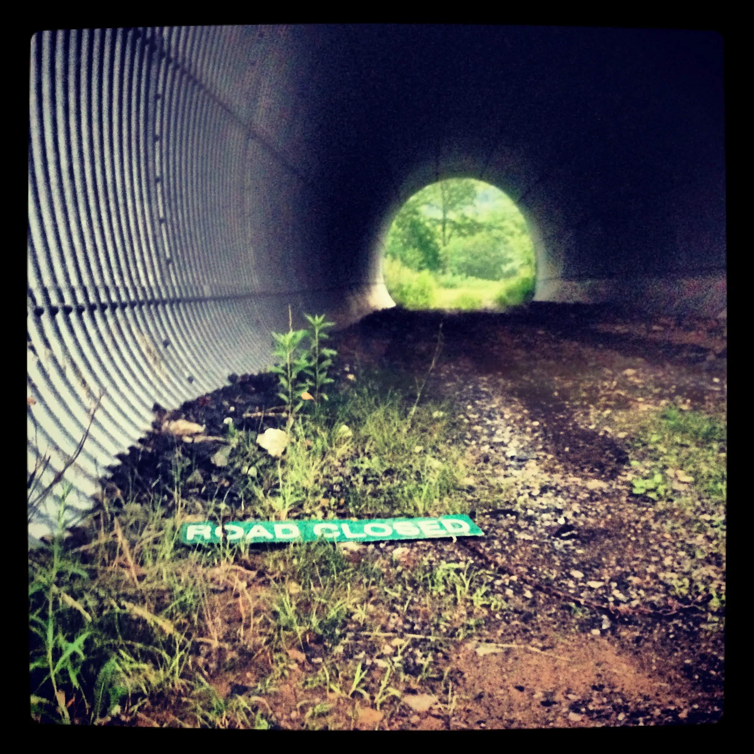 A corrugated tunnel opens toward green trees, with a small green sign reading “Road Closed” on the ground.