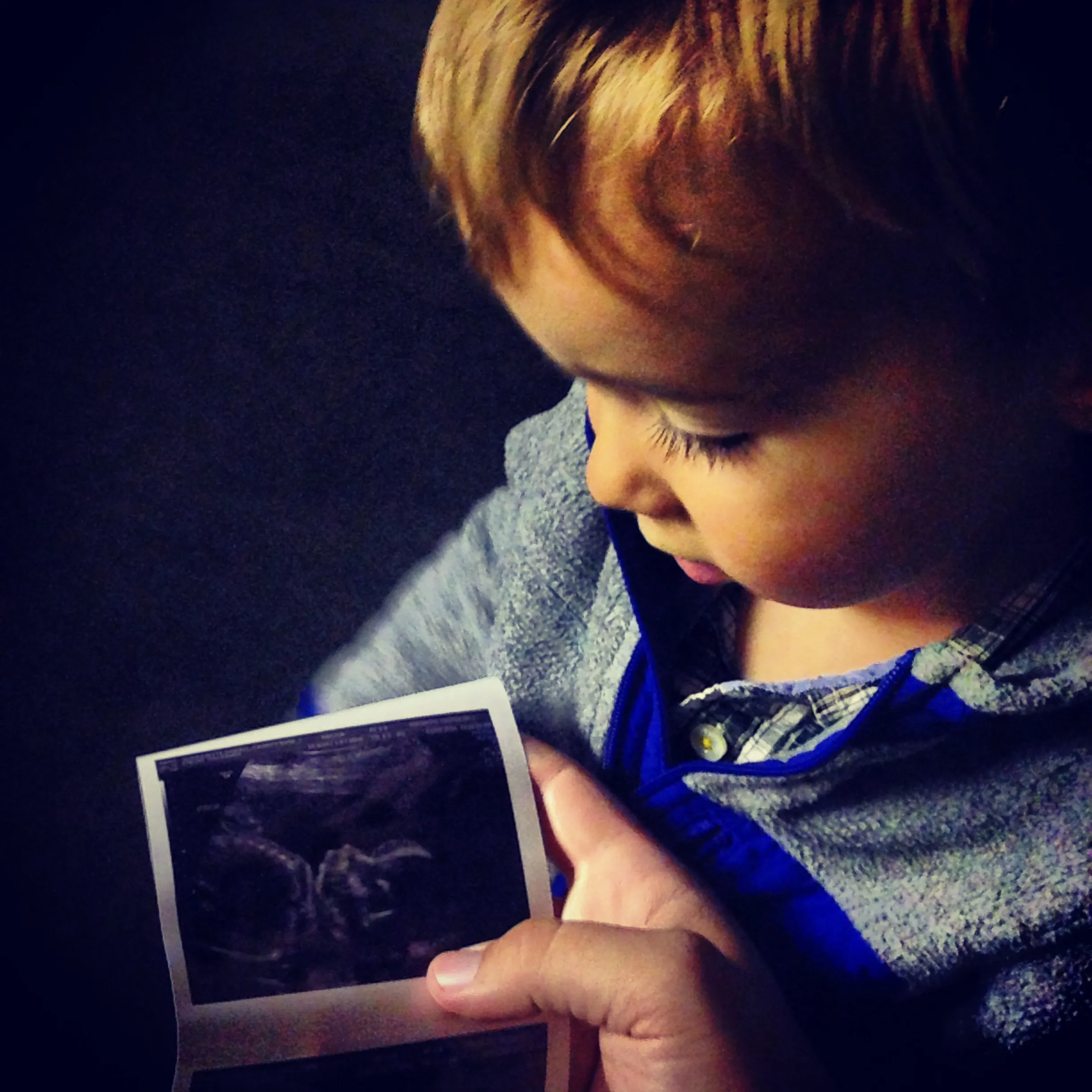 A young child looks down at an ultrasound print held in an adult hand.