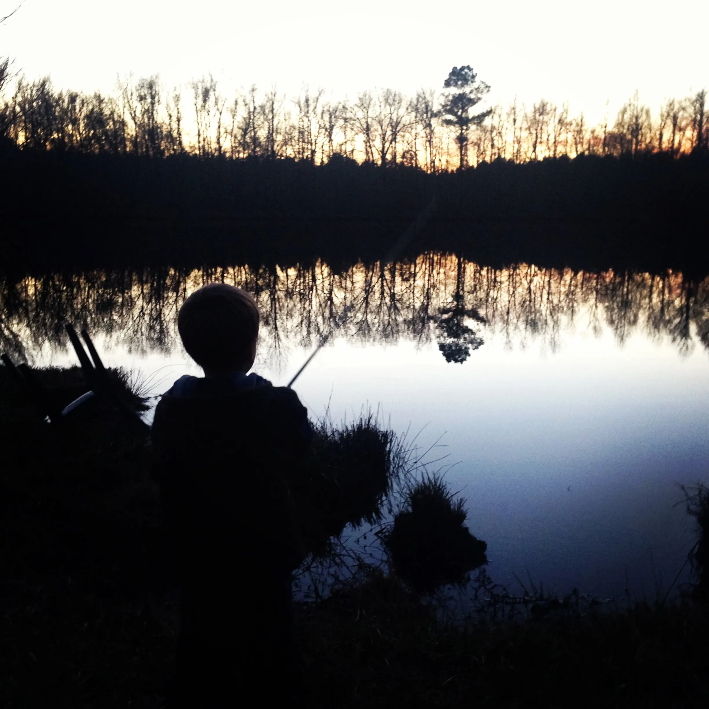 A silhouetted child holding a fishing rod stands beside a calm pond that reflects bare trees at sunset.