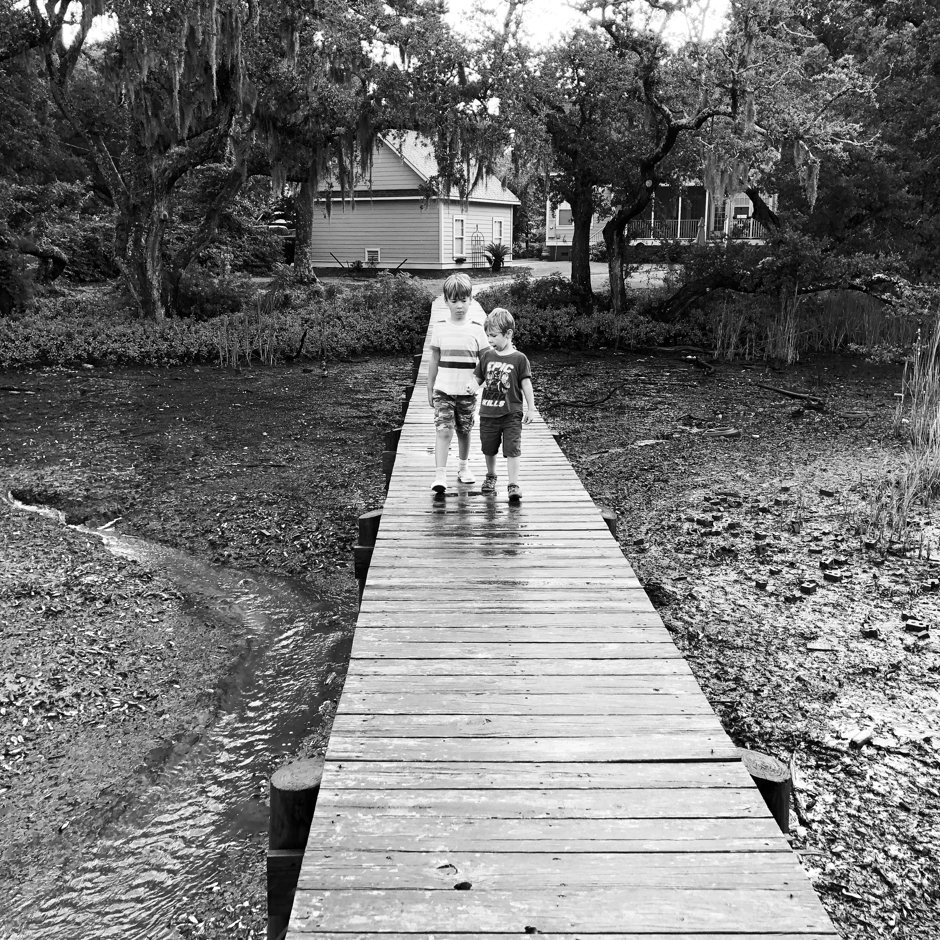 Black-and-white photograph of two children walking toward the camera on a narrow wet wooden boardwalk across muddy ground, with trees and houses behind them.