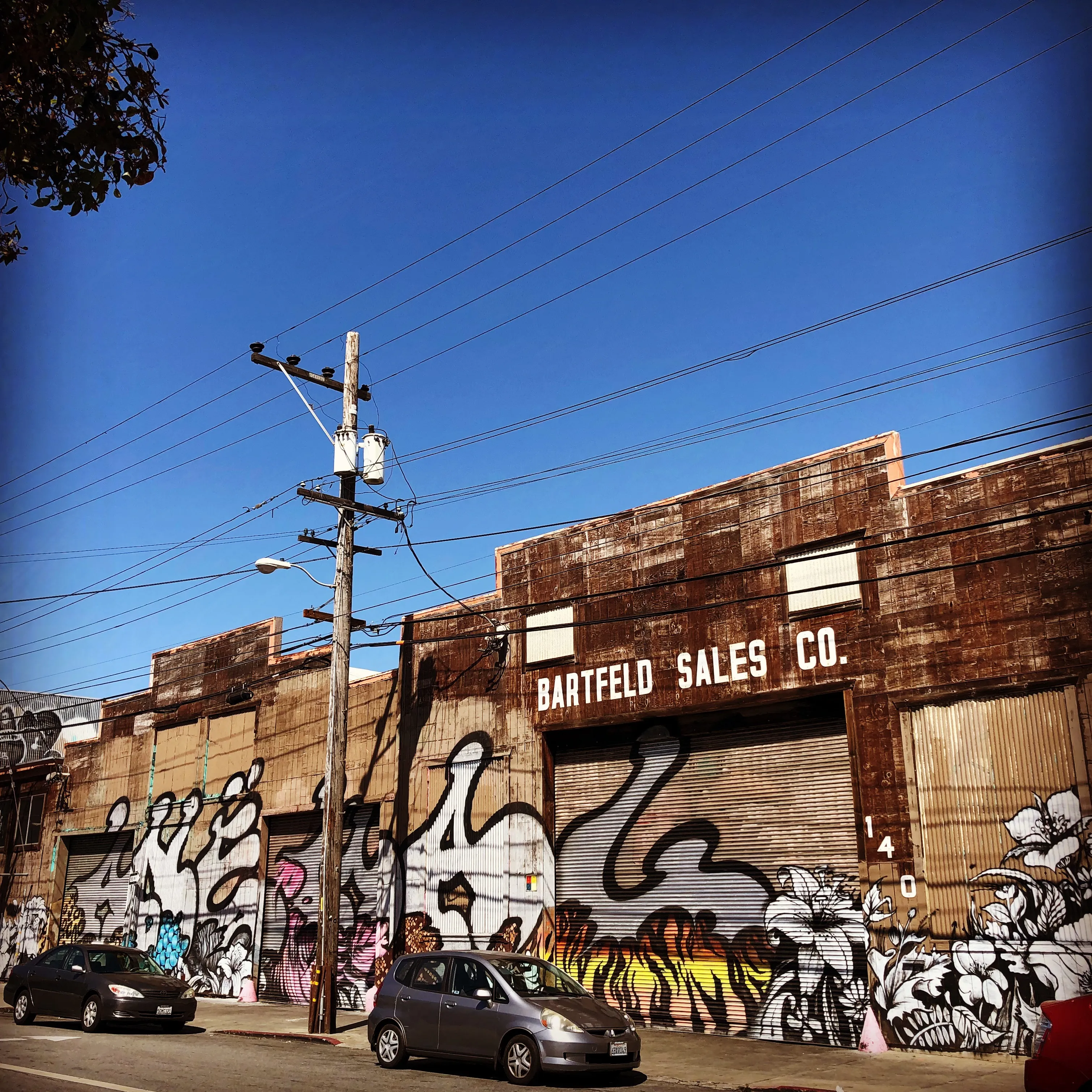 Street view of a low brick industrial building covered in black-and-white graffiti, parked cars, and overhead utility wires.