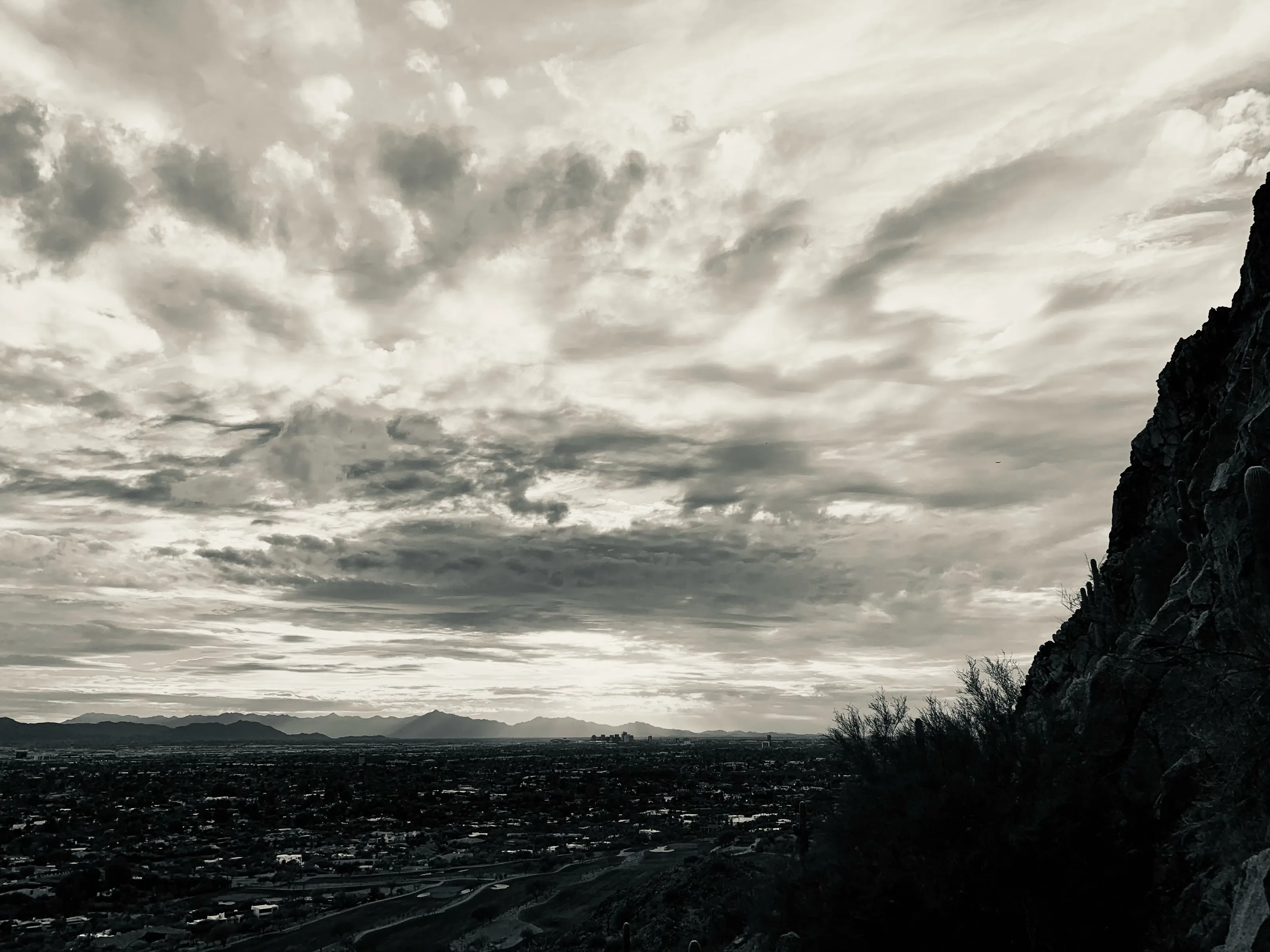 A black-and-white view from a rocky hillside over a city and distant mountains beneath dramatic clouds.