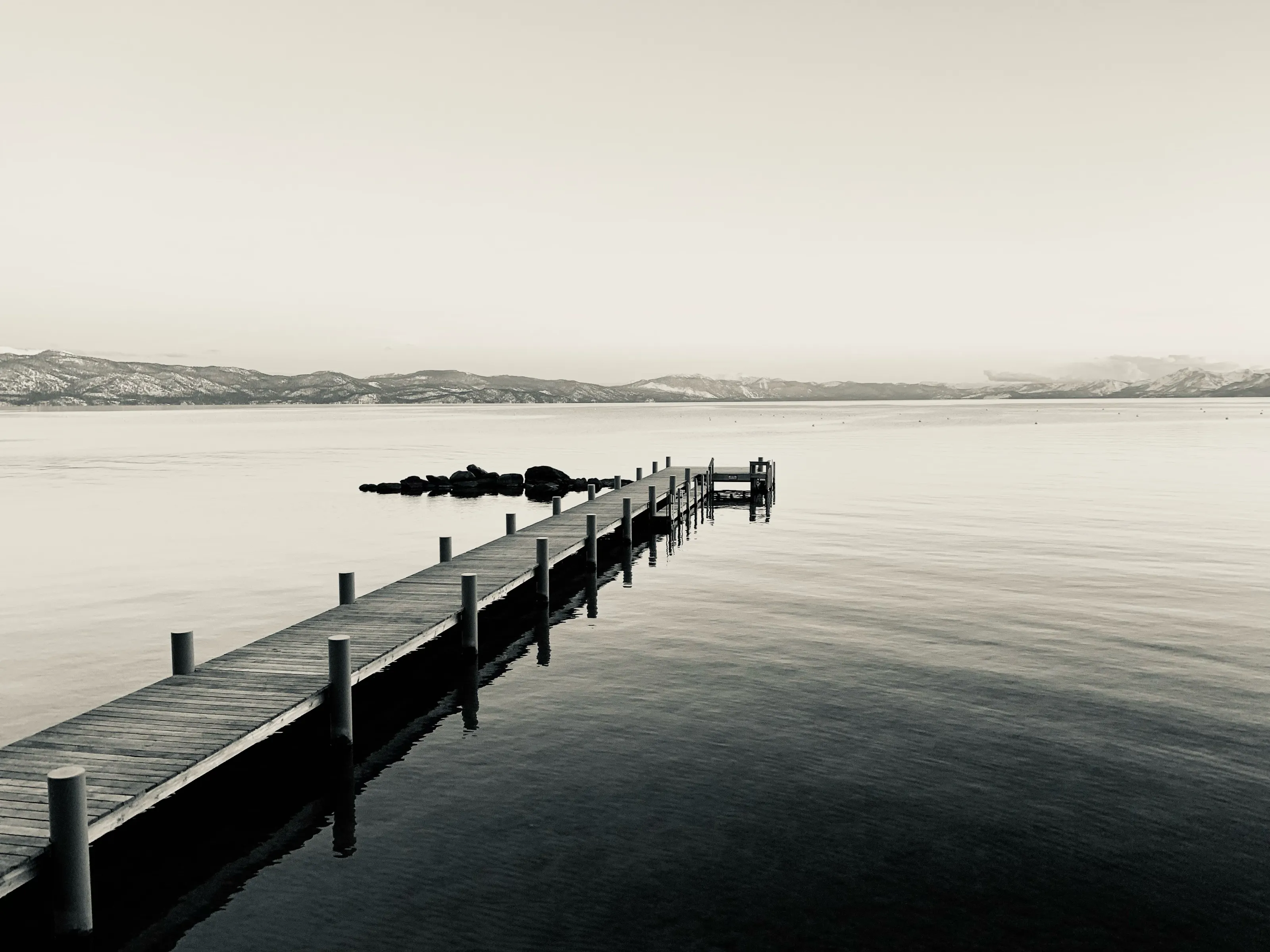A wooden dock projects into calm water toward low distant mountains in a muted monochrome landscape.