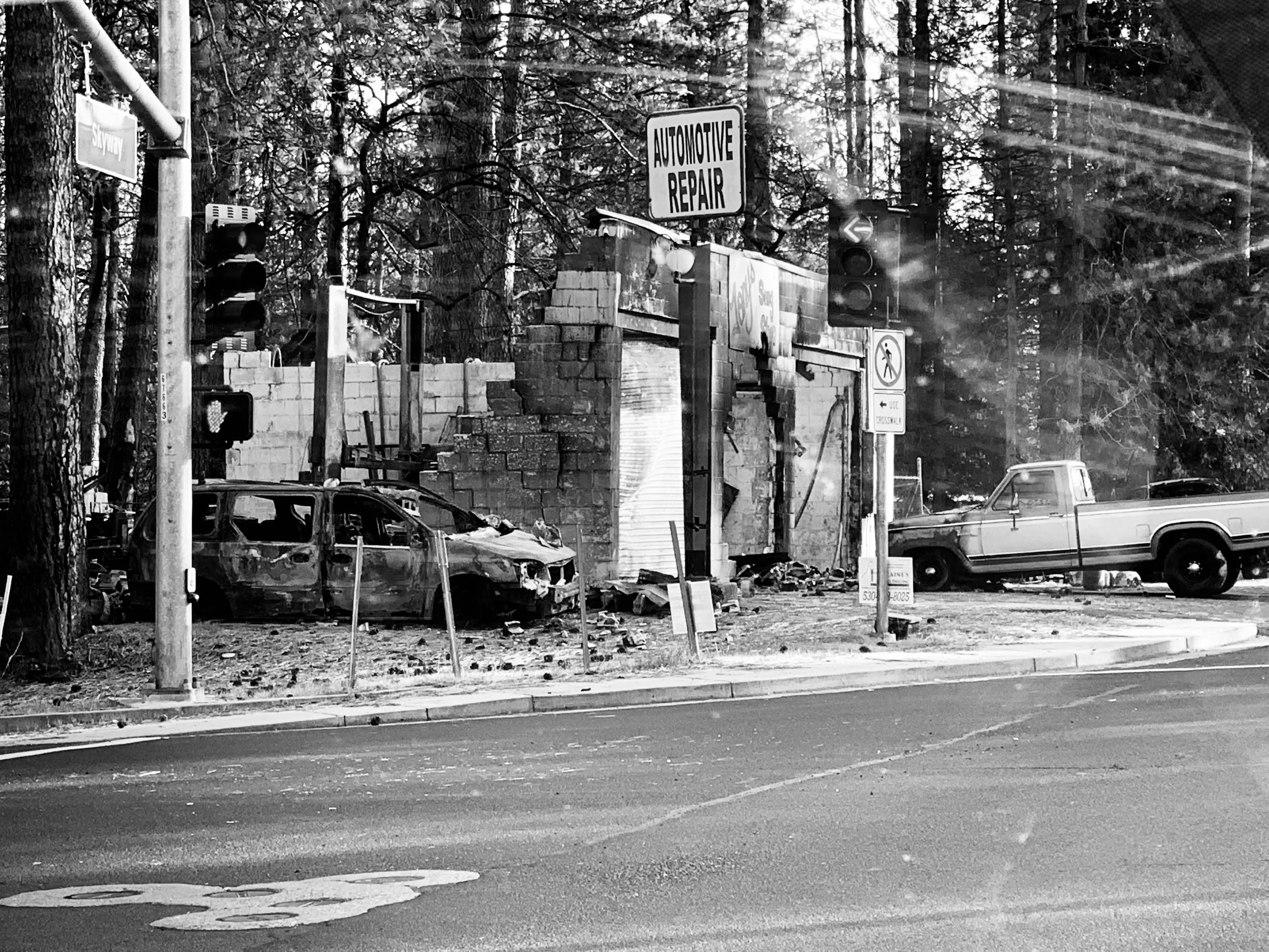 Black-and-white street scene showing a damaged automotive repair building, a burned car, traffic lights, and a pickup truck.