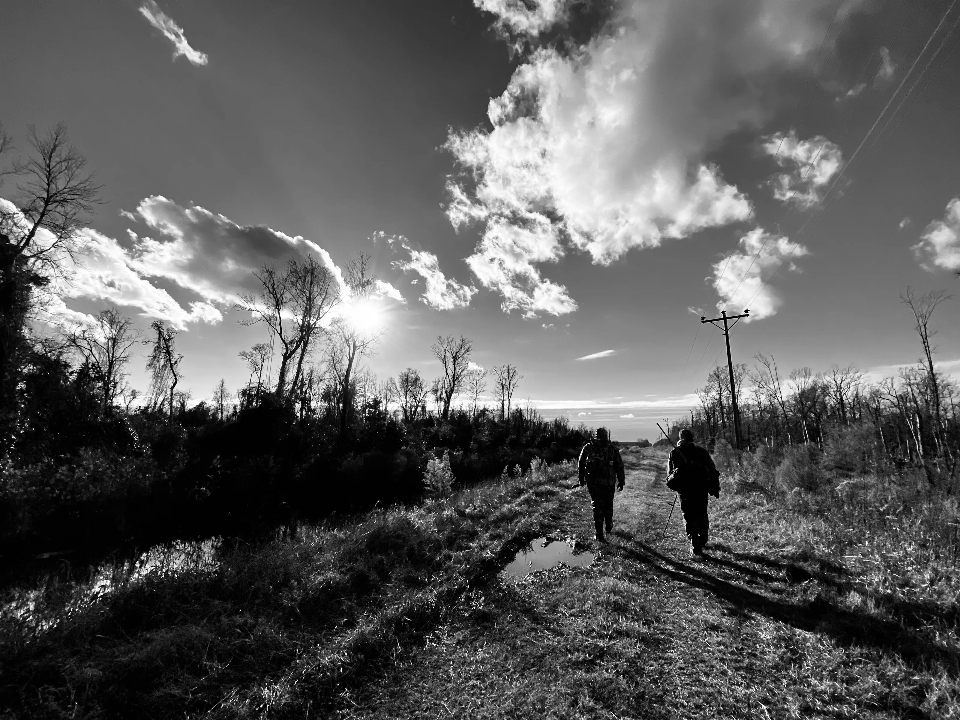 Black-and-white photograph of two people walking away along a muddy road with puddles, utility poles, bare trees, and bright clouds overhead.